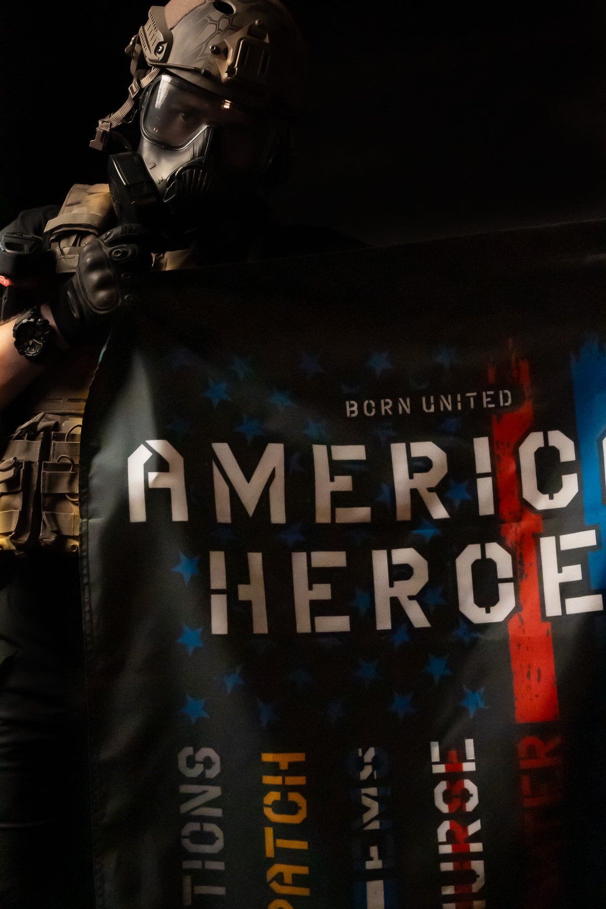 A person wearing tactical gear holds a Born United Private Label American Heroes Flag. The blue and red design, emblazoned with BORN UNITED AMERICAN HEROES and featuring stars and WATCH, JUDGE, and FATHER, boldly honors military valor against a dark backdrop.