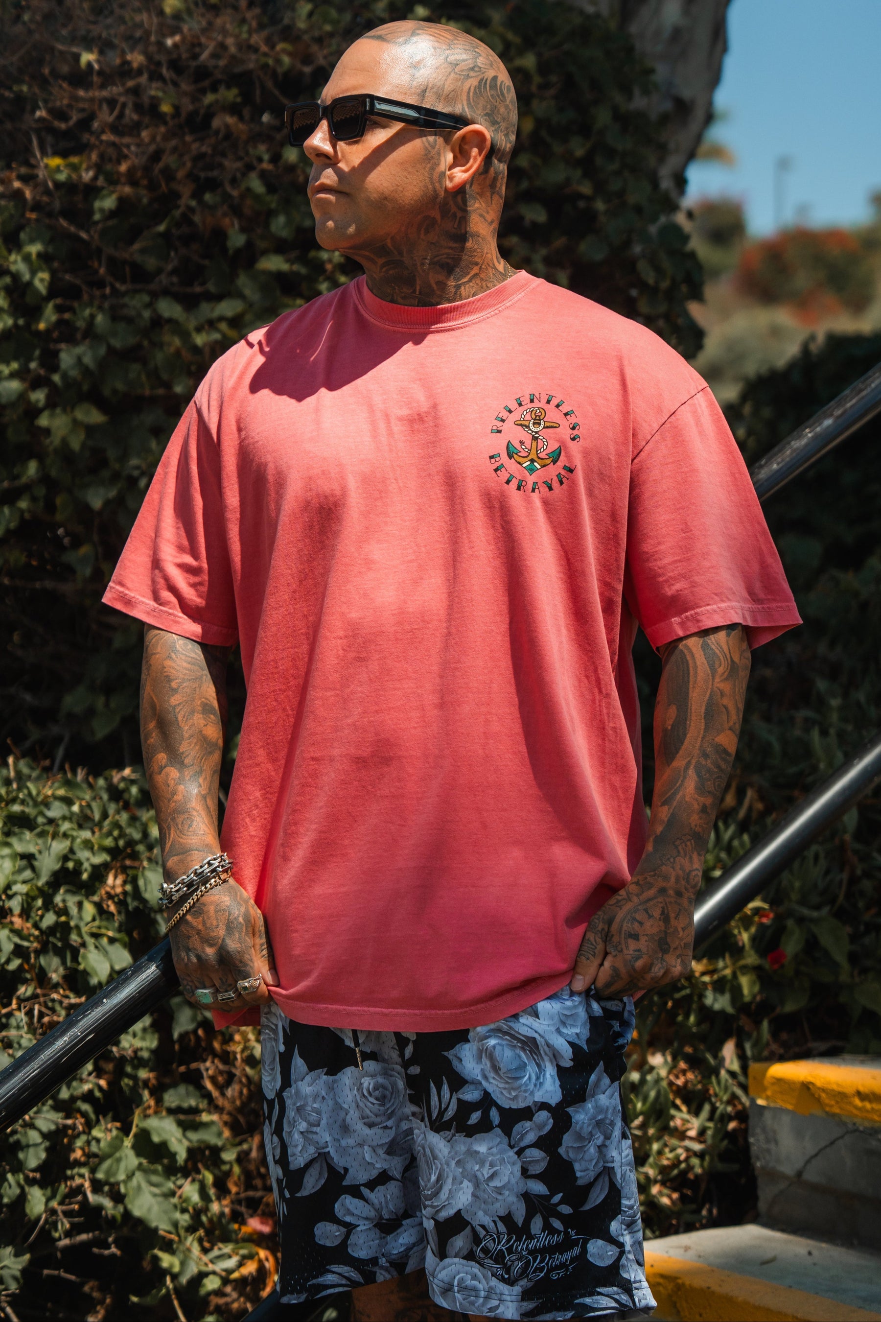 A tattooed man in sunglasses and the Relentless Betrayal Into the Storm Premium Watermelon T-Shirt, paired with black-and-white floral shorts, stands outdoors near stairs amid greenery.