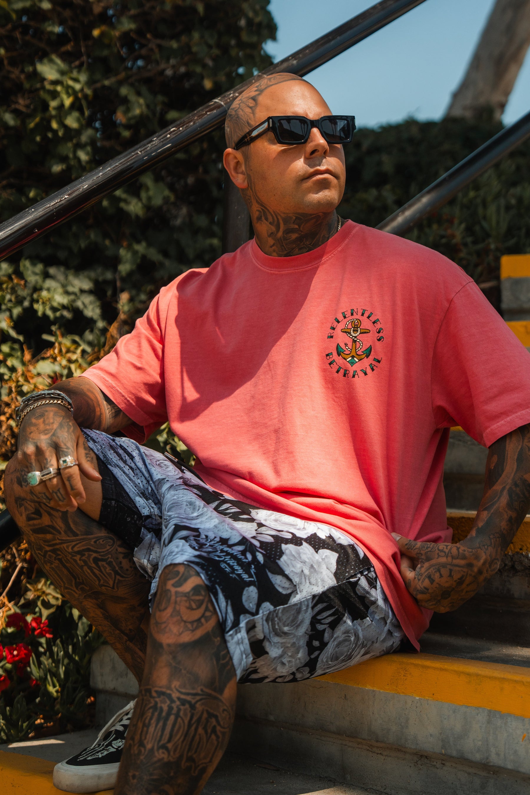A tattooed man sits on outdoor steps in black sunglasses and a Relentless Betrayal Into the Storm Premium Watermelon T-Shirt. Sunlight shines on him, and greenery is visible in the background.