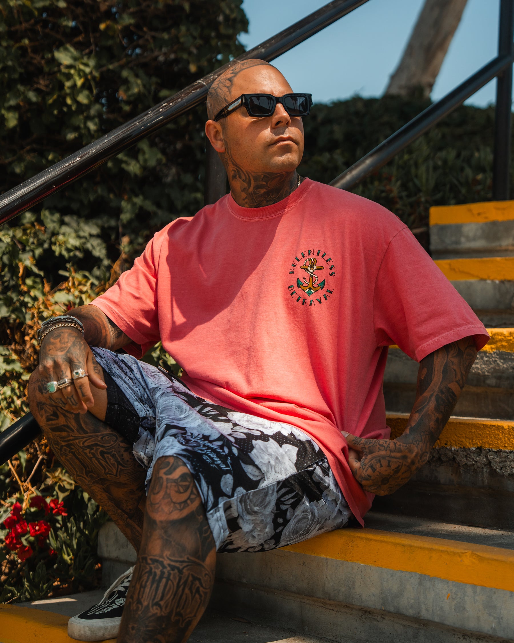 A tattooed man in sunglasses, a pink T-shirt, and Relentless Betrayals Premium Mesh Shorts | 5- Beauty in Darkness sits on outdoor stairs amidst greenery, sunlight creating dramatic shadows around him.