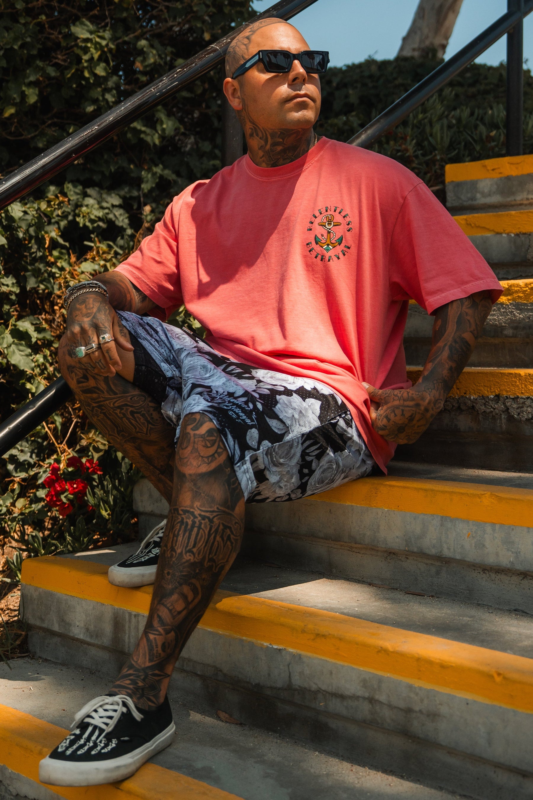A tattooed man in sunglasses and patterned shorts sits on outdoor steps, wearing the Relentless Betrayal Into the Storm Premium Watermelon T-Shirt, made of jersey cotton, as he looks off to the side on a sunny day.