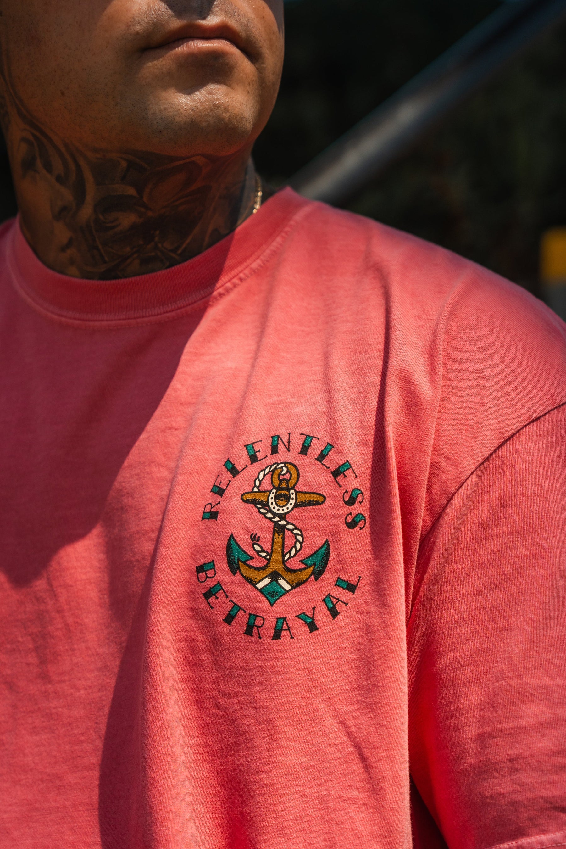 A person wears the Relentless Betrayal Into the Storm Premium Watermelon T-Shirt, featuring an anchor graphic and branding on the chest. Neck tattoos are visible; their face is partially out of frame.