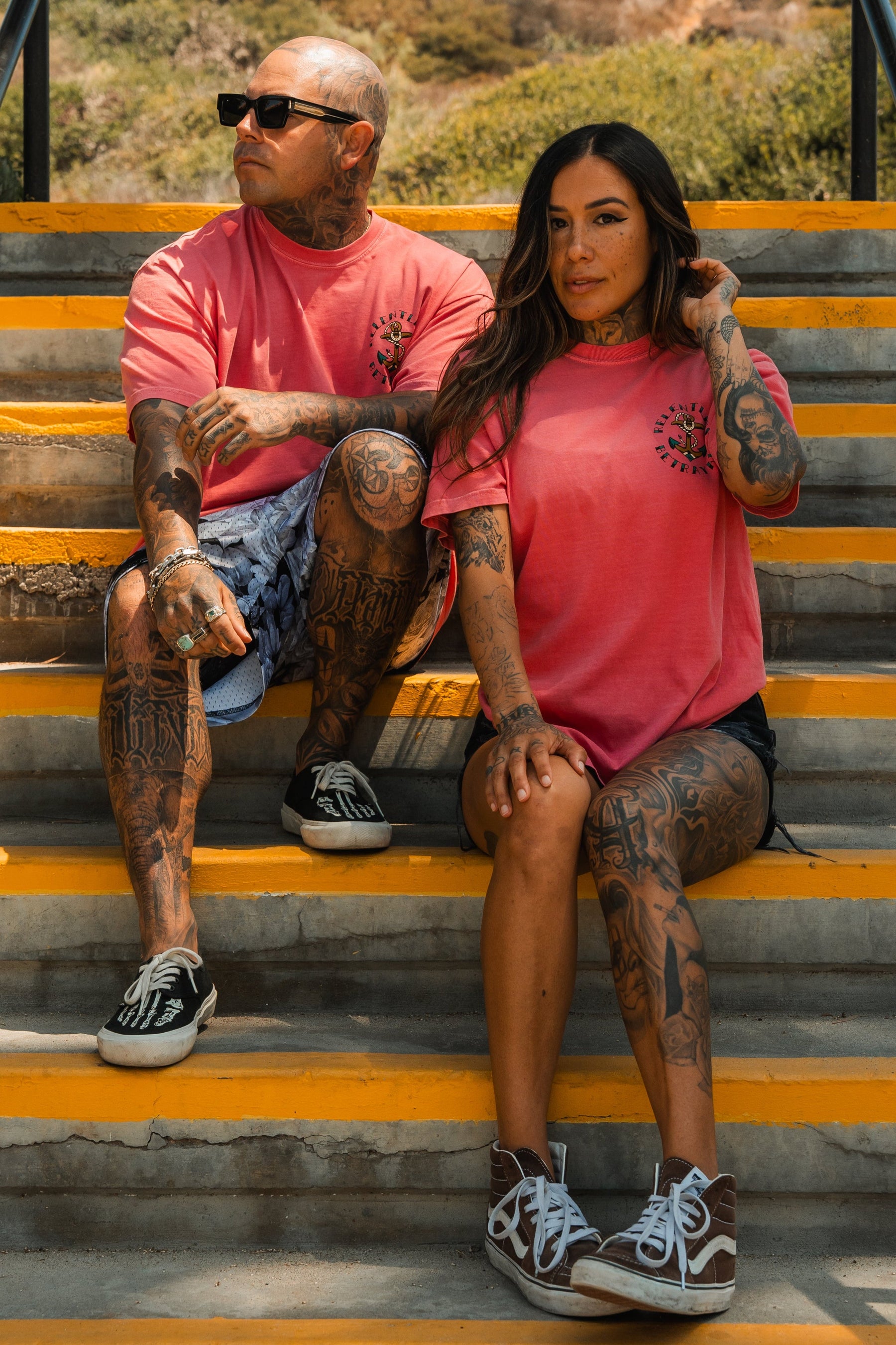 A tattooed man and woman sit on outdoor stairs amid greenery, both in pink unisex Relentless Betrayal shirts—he sports the Into the Storm Premium Watermelon T-Shirt. Relaxed and stylish in shorts, they enjoy each others company.