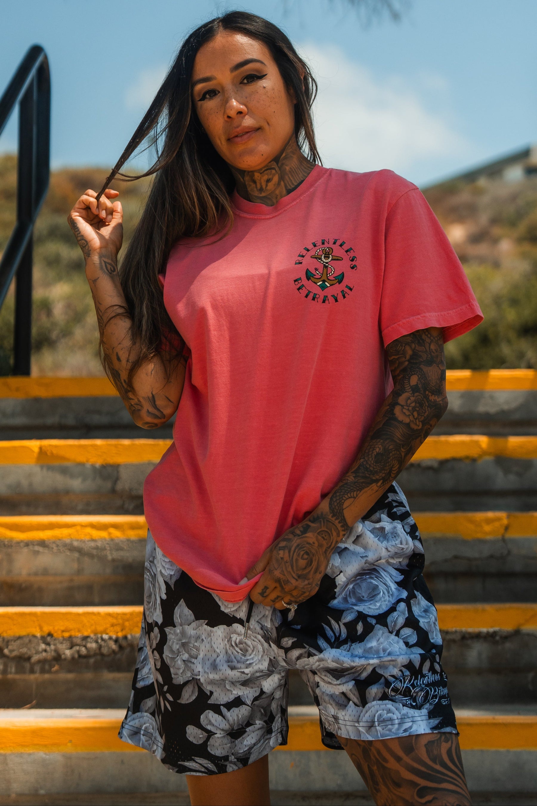 A tattooed woman wearing the Relentless Betrayal Into the Storm Premium Watermelon T-Shirt and black-and-white floral shorts stands on yellow-striped outdoor stairs, holding her long hair, with trees and sky behind her.