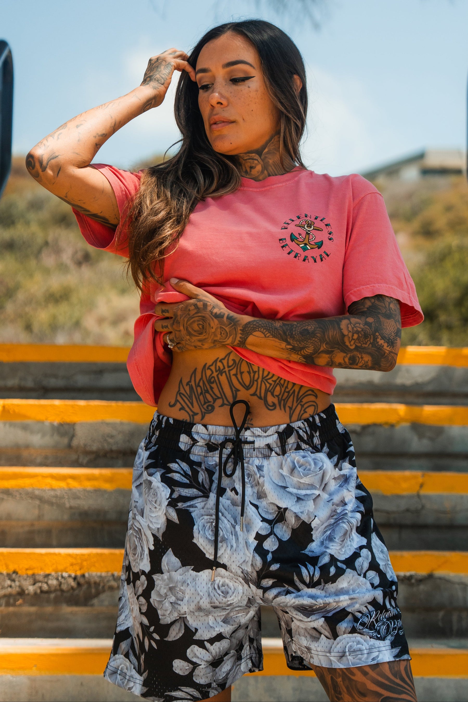 A tattooed woman poses confidently outdoors on steps, wearing the Relentless Betrayal Into the Storm Premium Watermelon T-Shirt and floral shorts. She touches her hair, with one hand on her head and the other near her waist.