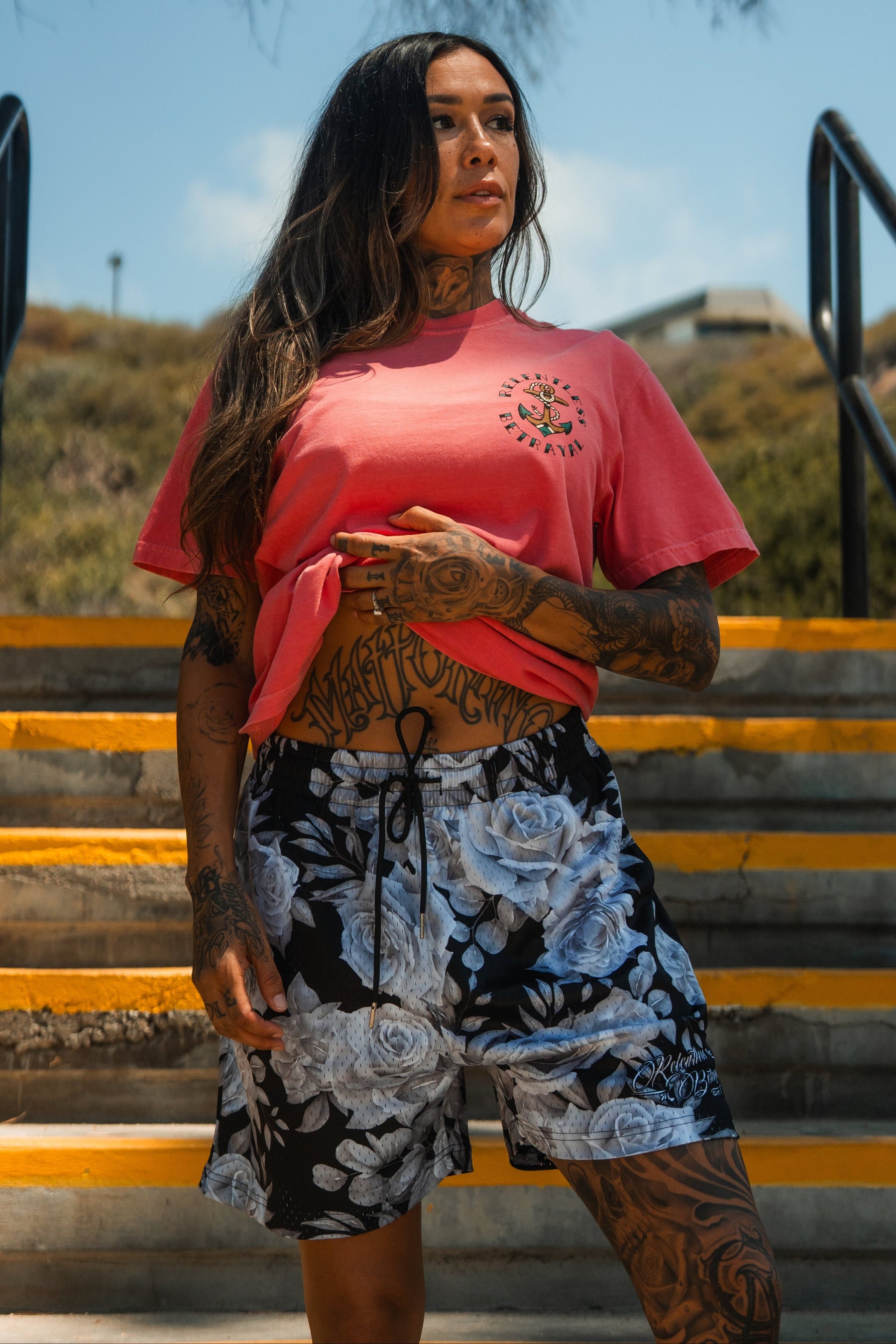 A woman with long hair and tattoos stands on outdoor steps, wearing the Relentless Betrayal Into the Storm Premium Watermelon T-Shirt tied at the waist and black floral shorts. Trees and a building can be seen in the background.