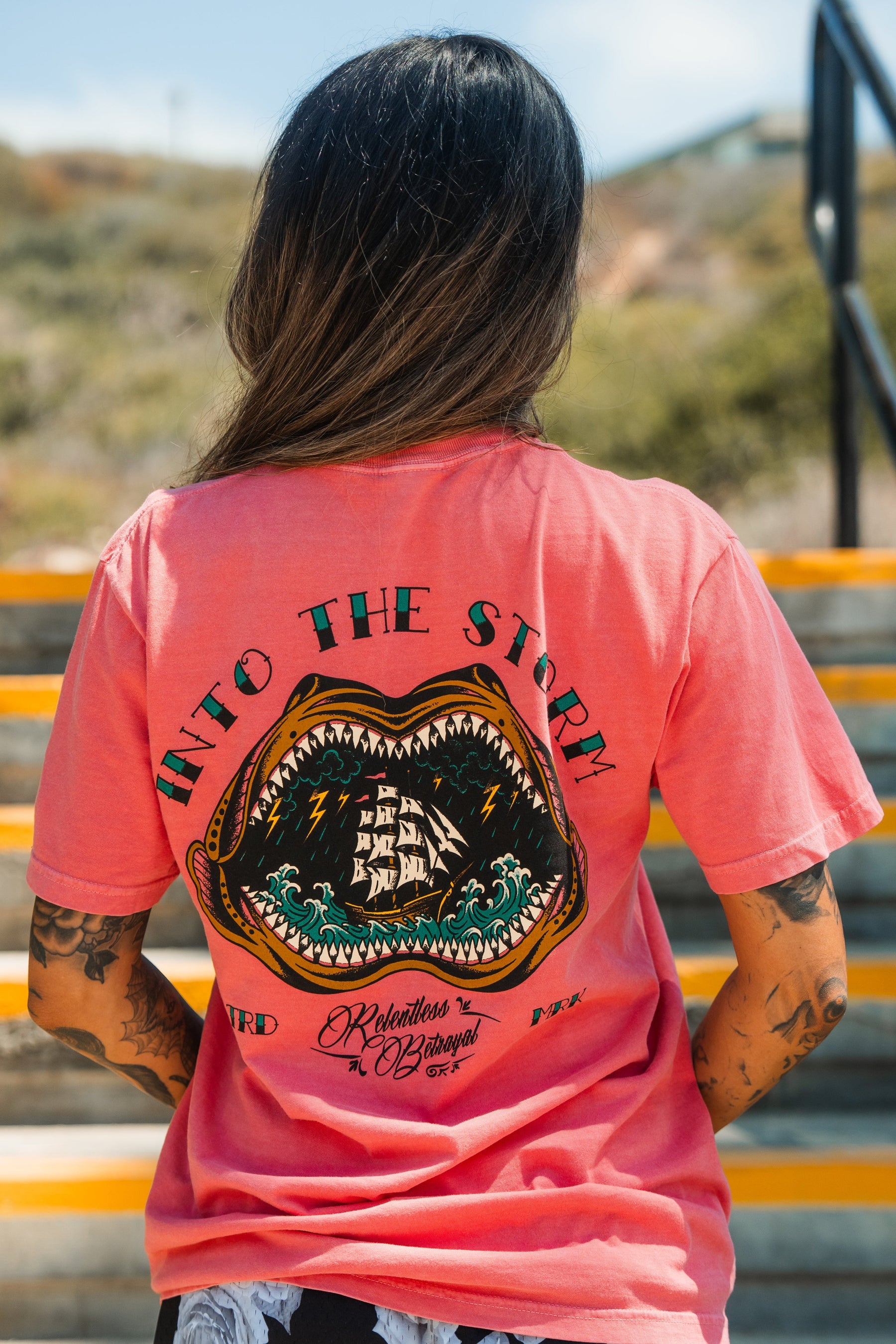 A tattooed person wears the Relentless Betrayal Into the Storm Premium Watermelon T-Shirt, featuring a ship in waves inside a shark mouth and “INTO THE STORM” on the back, while standing on outdoor stairs.