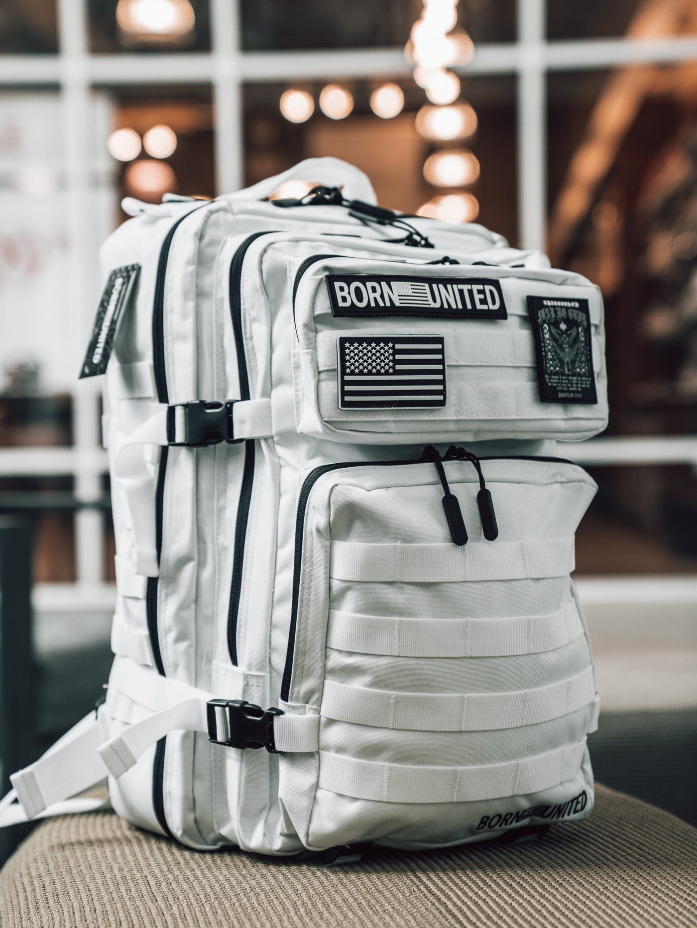 The Standard Tac-Pacs Ultimate Bundle by Born United Private Label, a white tactical backpack with black straps, multiple zippered compartments, and BORN UNITED and American flag patches, rests on a beige cushioned surface indoors.