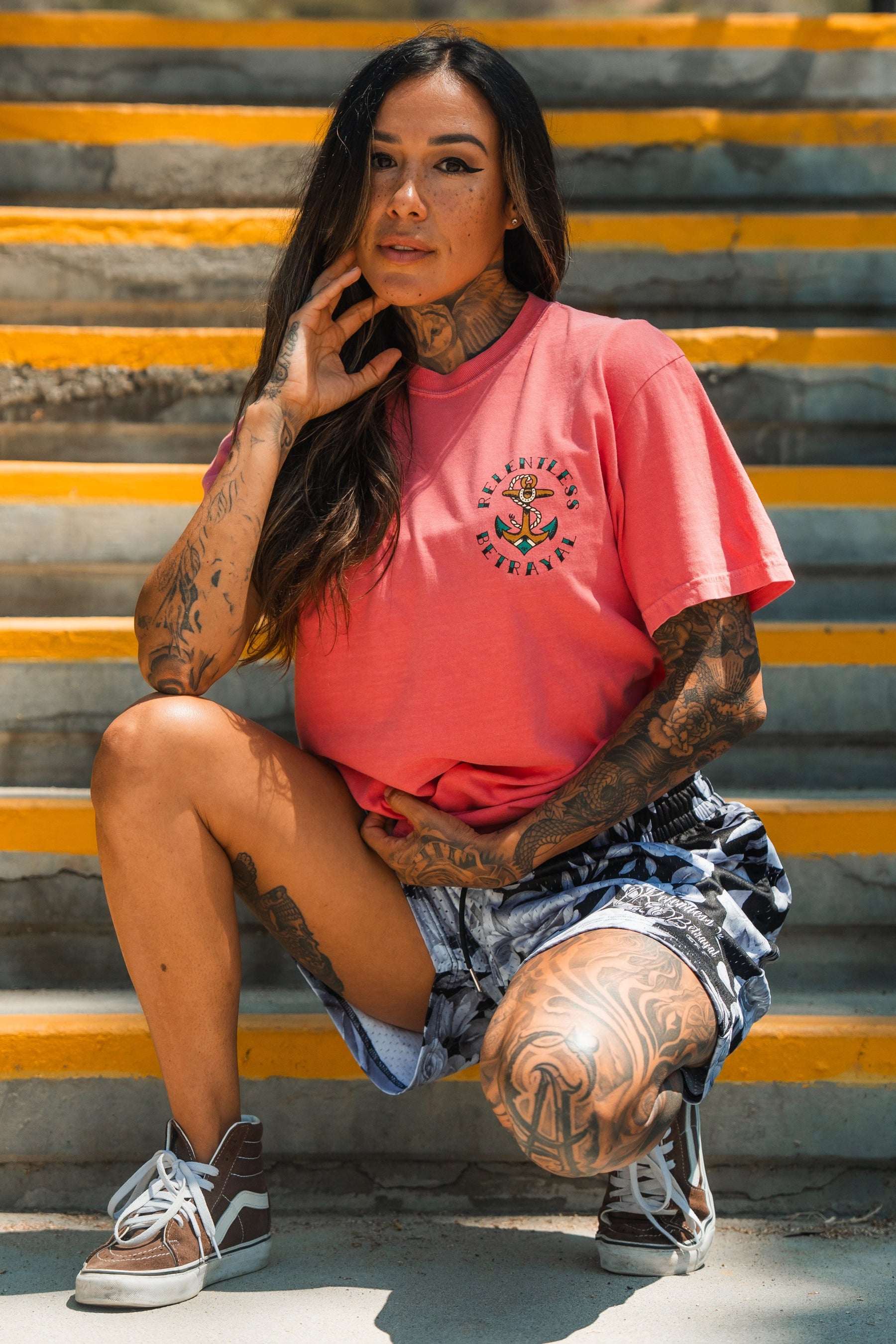 A woman with long brown hair and tattoos poses confidently on yellow-striped concrete steps, wearing the Relentless Betrayal Into the Storm Premium Watermelon T-Shirt made from jersey cotton.