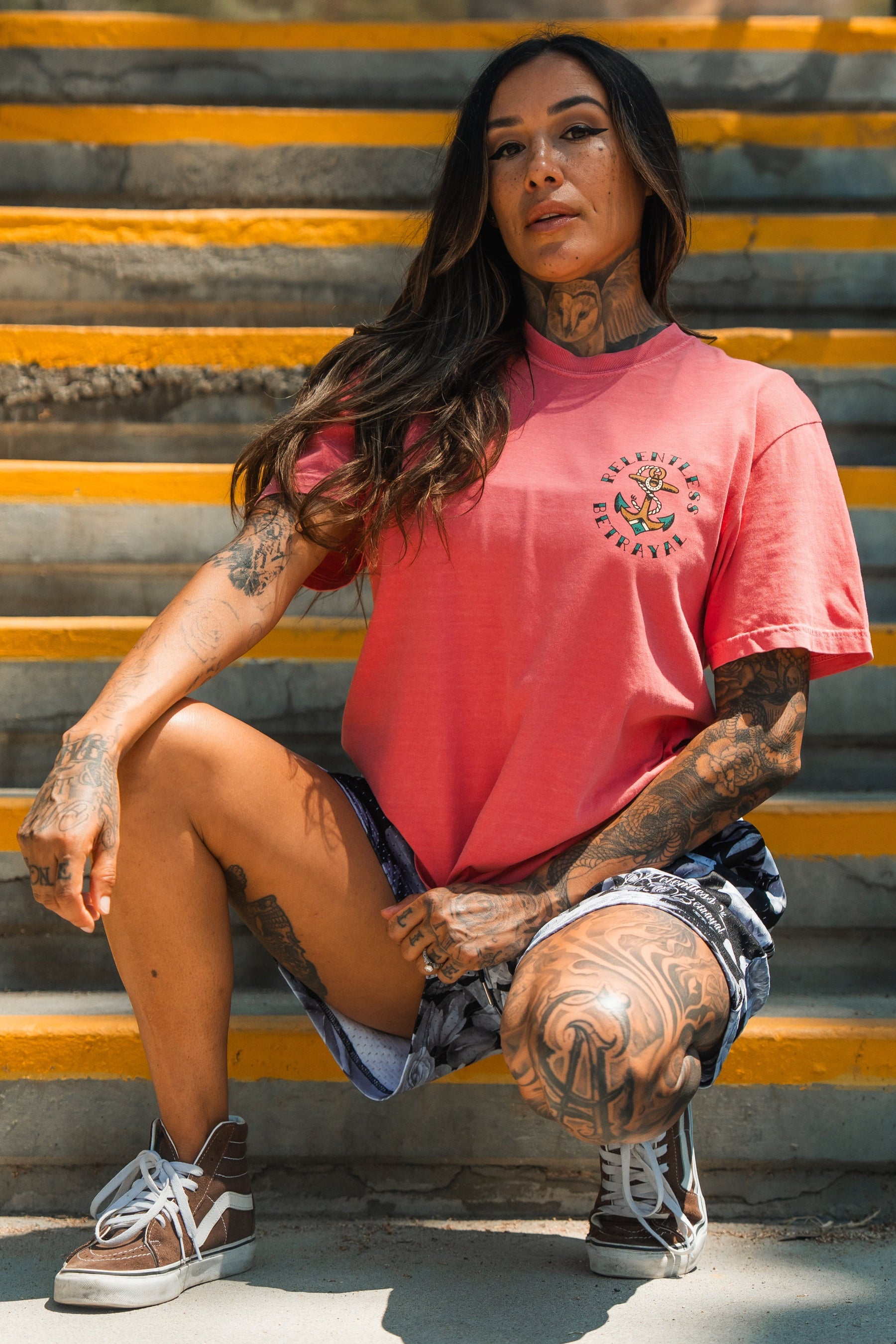 A tattooed woman with long dark hair kneels on outdoor yellow-edged steps, confidently wearing the Relentless Betrayal Into the Storm Premium Watermelon T-Shirt, paired with shorts and sneakers.