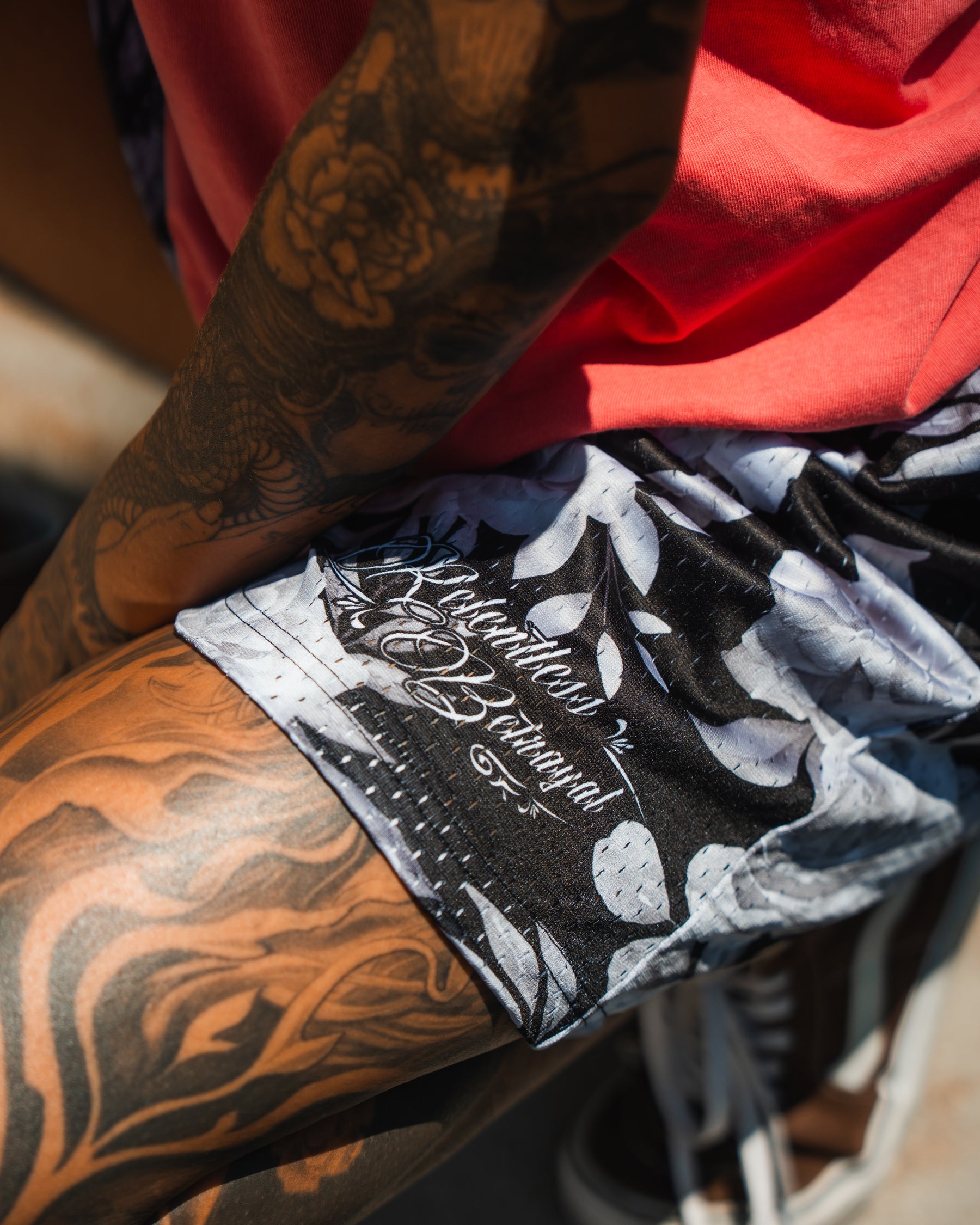 A close-up of a tattooed arm and leg wearing Relentless Betrayals Premium Mesh Shorts | 5\- Beauty in Darkness with cursive writing, paired with a coral shirt. A blurred background hints at an outdoor setting.