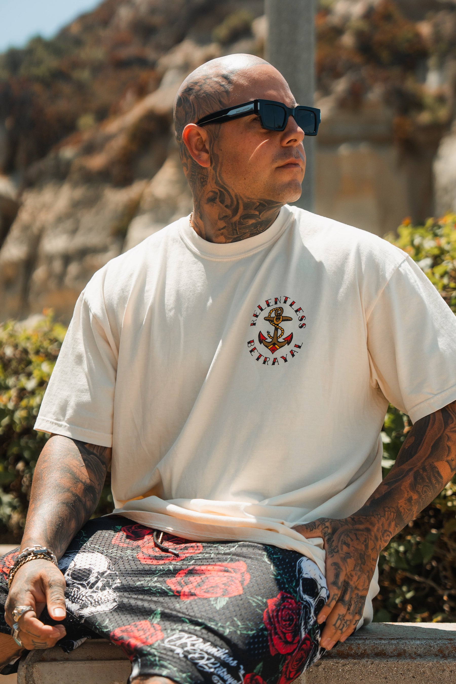 A tattooed man in sunglasses wears the Relentless Betrayal Into the Storm Premium Ivory T-Shirt with a small logo and patterned shorts, sitting outdoors on a sunny day amid greenery and rocky hills.