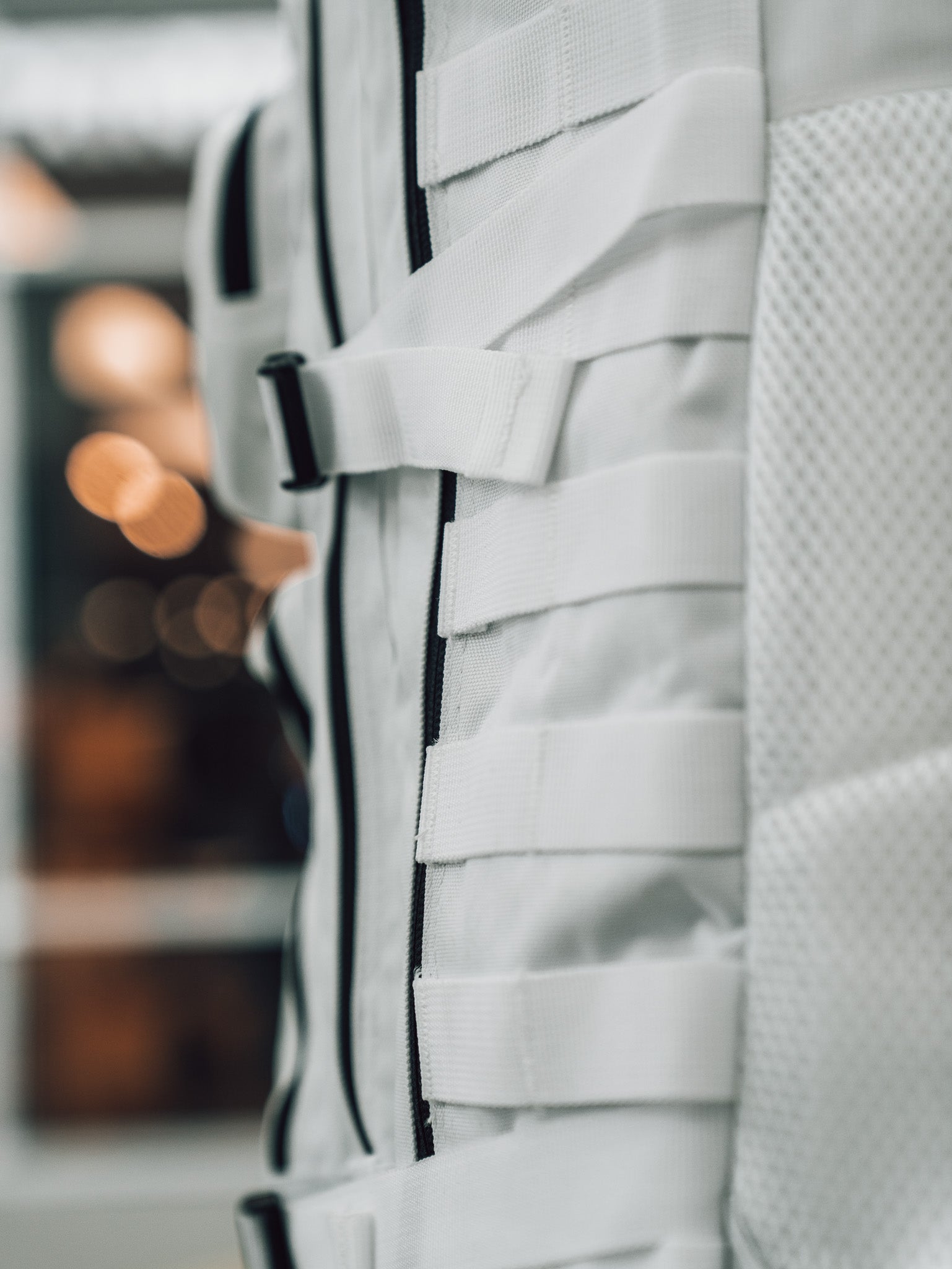 Close-up of the Born United Private Label Storm Trooper Tac-Pac - 45L, a white fabric bag with horizontal straps and mesh texture, inspired by Star Wars, set against a blurred background with warm lights.