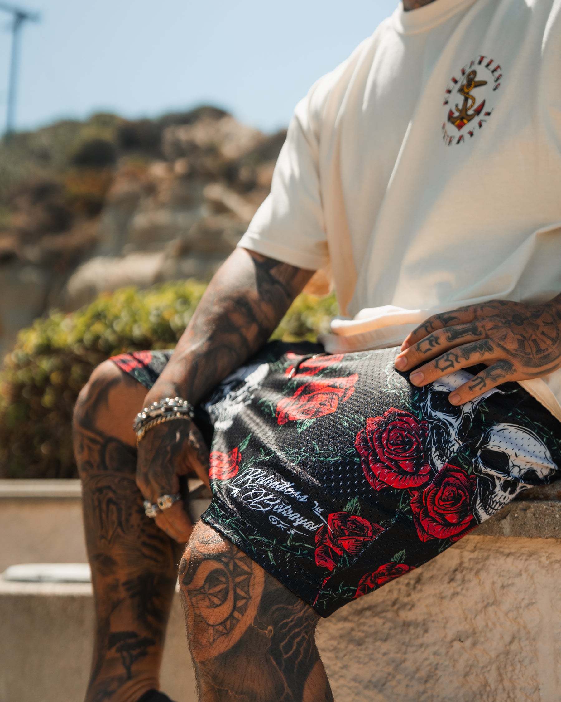 A tattooed person sits outdoors in a white T-shirt and Relentless Betrayal Premium Mesh Shorts | 5 - Thorn Amongst Roses, featuring red roses and skulls. The sunny scene includes blurred greenery and rocky hills in the background.