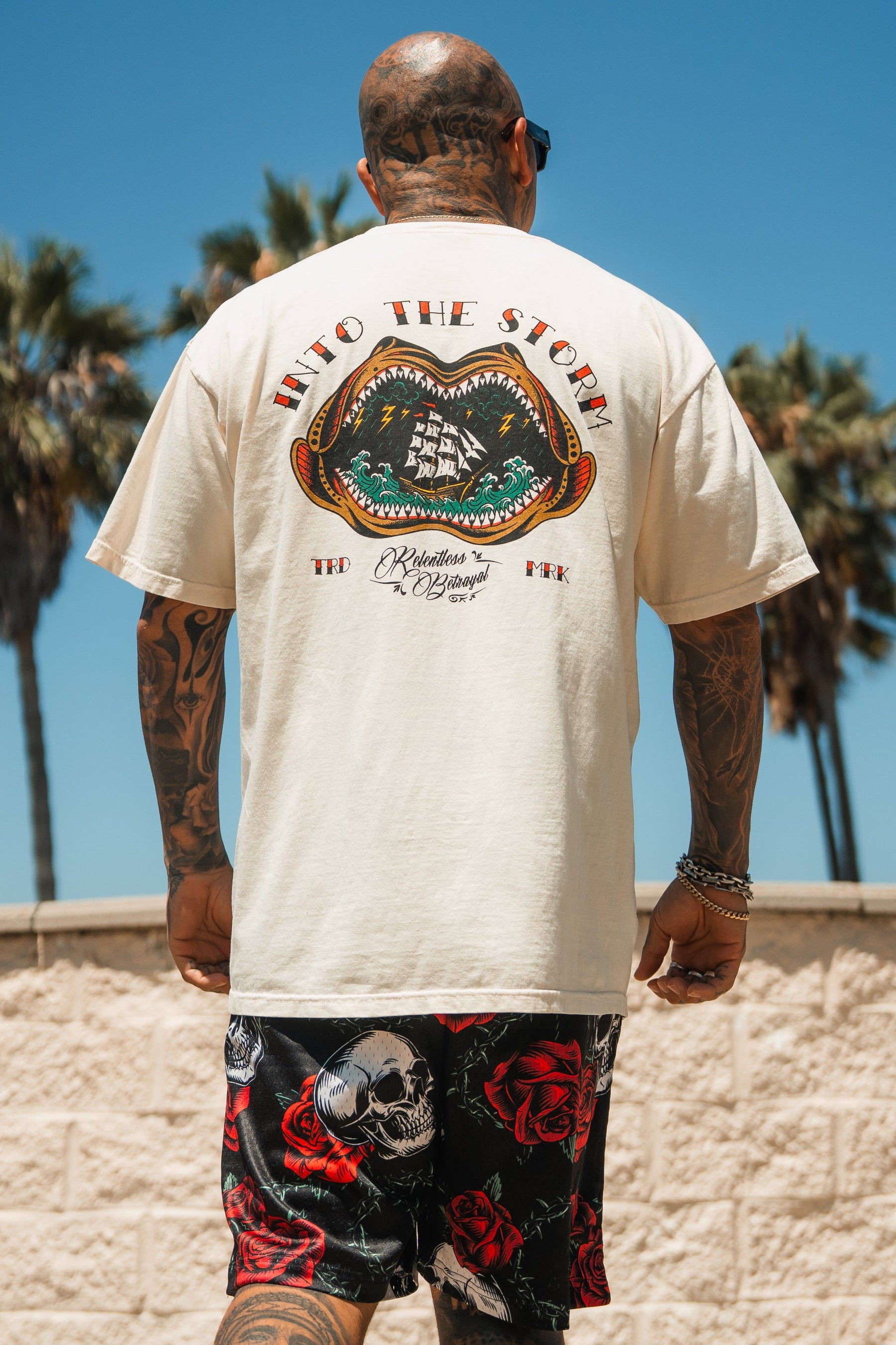 A tattooed man stands outdoors under palm trees and blue sky, wearing the Relentless Betrayal Into the Storm Premium Ivory T-Shirt with a ship graphic, plus black shorts patterned with skulls and red roses.