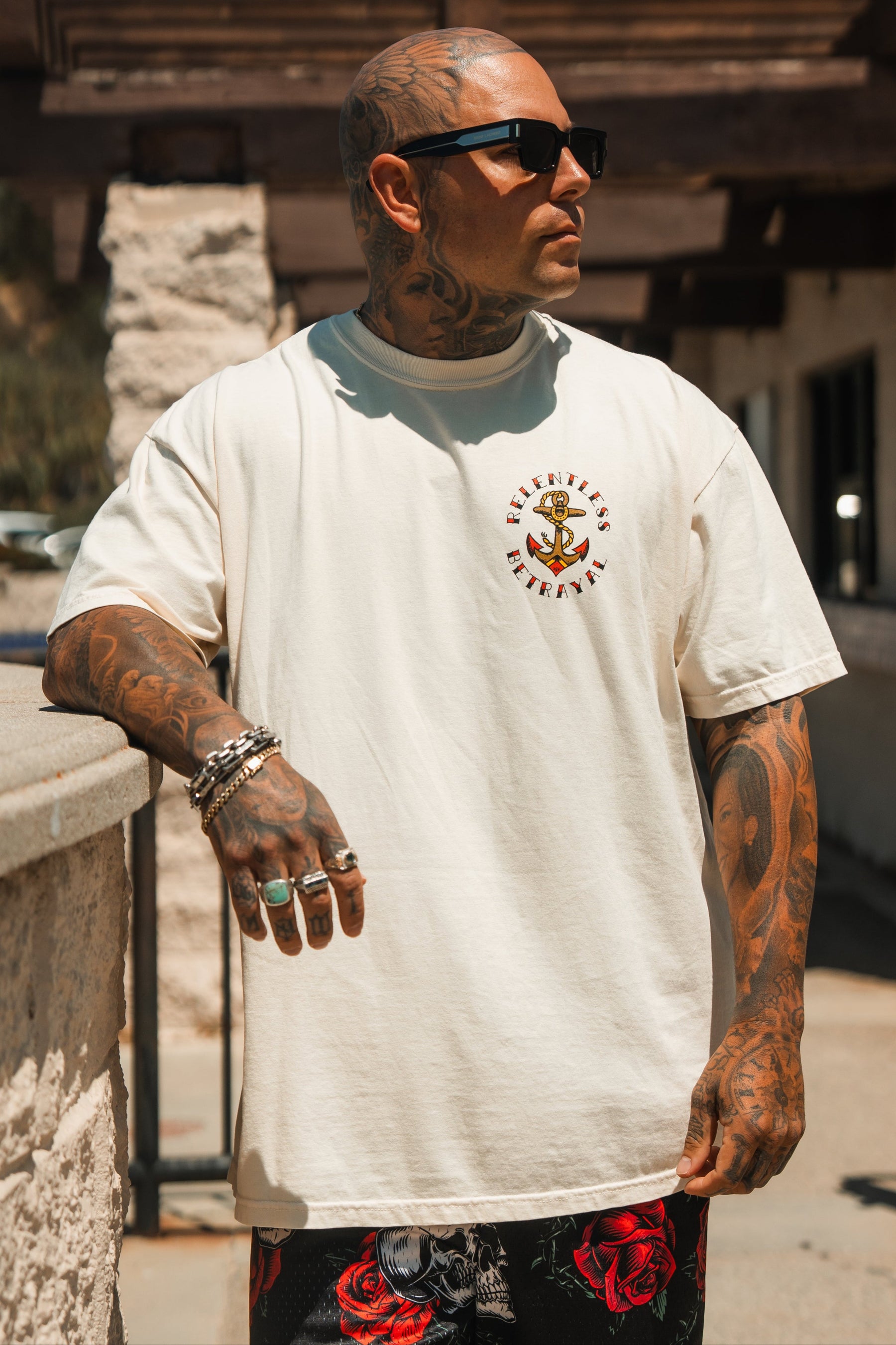 A tattooed man in sunglasses wears the Relentless Betrayal Into the Storm Premium Ivory T-Shirt and black shorts with red roses, standing outdoors by a stone wall and looking to the side.