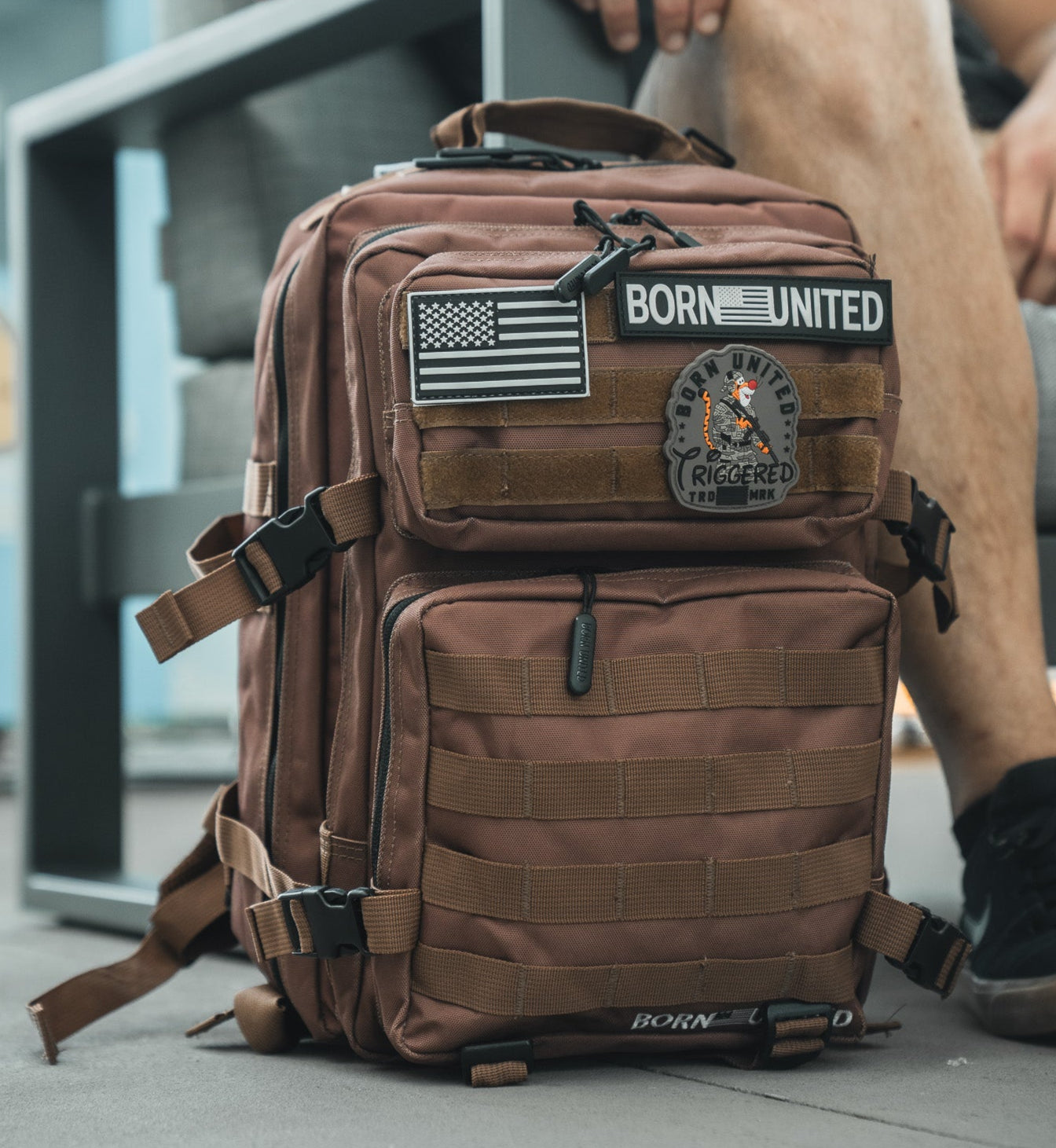 A Chocolate Tac-Pac - 25L by Born United Private Label, TSA-approved tactical backpack with multiple compartments and American flag, BORN UNITED, and TRIGGERED patches. Part of a person’s leg and shoe are visible beside the bag.