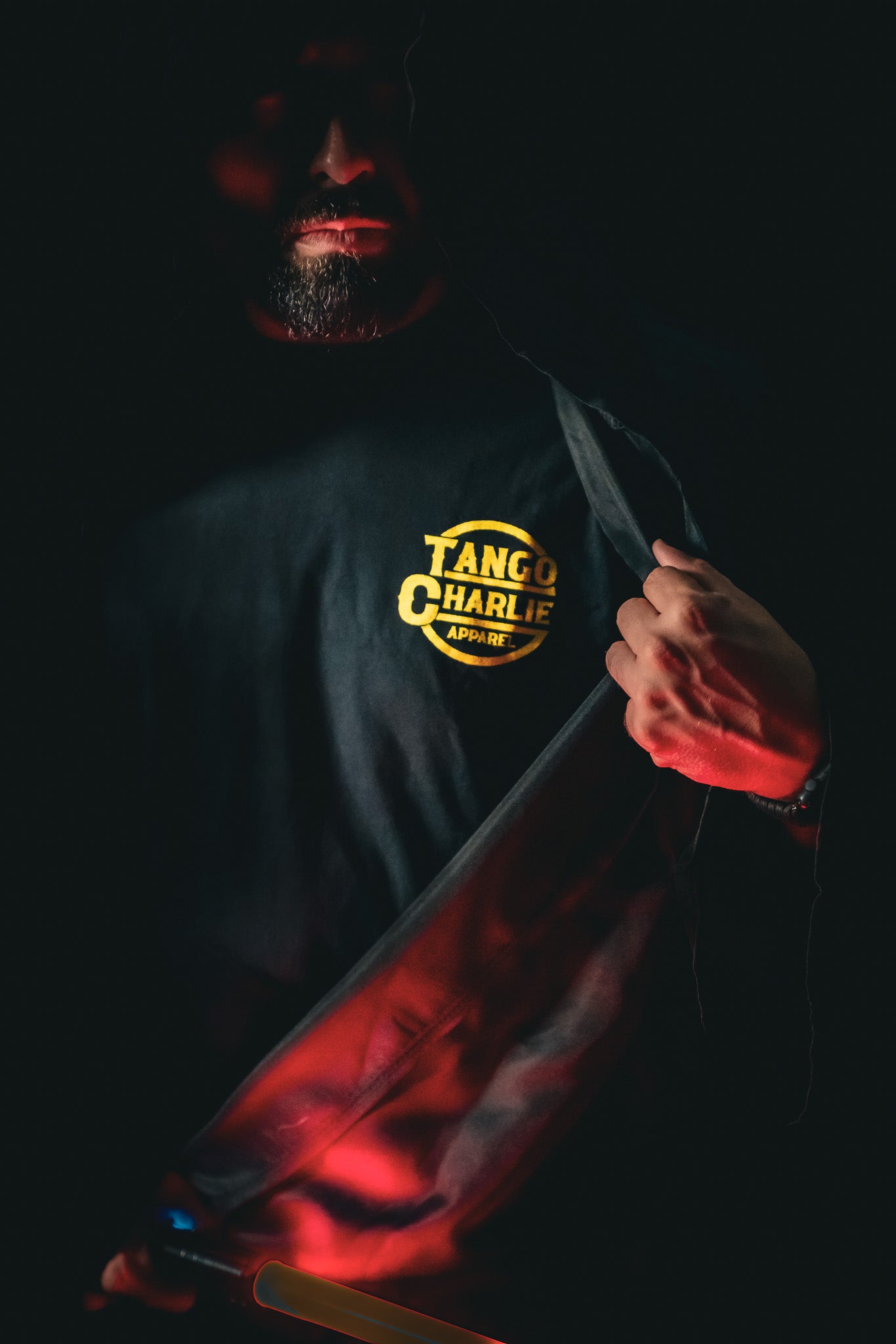 A person in shadow opens a dark coat, revealing a limited edition Lift Vader T-Shirt by Tango Charlie Apparel. A red light highlights their hand and shirt, creating a dramatic contrast against the darkness, hinting at their intense fitness journey.