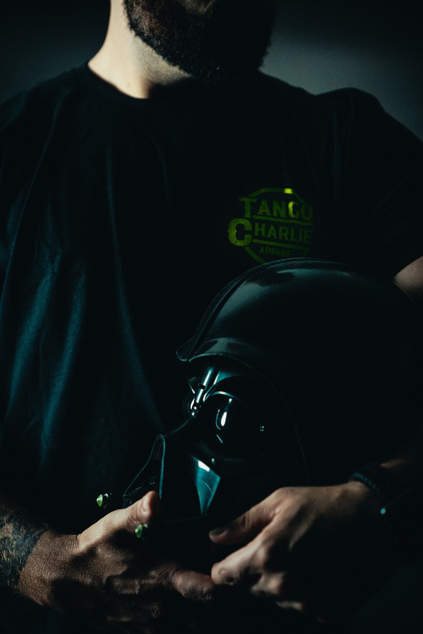 A bearded man in a dark Tango Charlie Apparel T-shirt cradles a limited edition, black helmet reminiscent of a sci-fi villains. The dim lighting enhances the mystery, like an exclusive Lift Vader fitness challenge.
