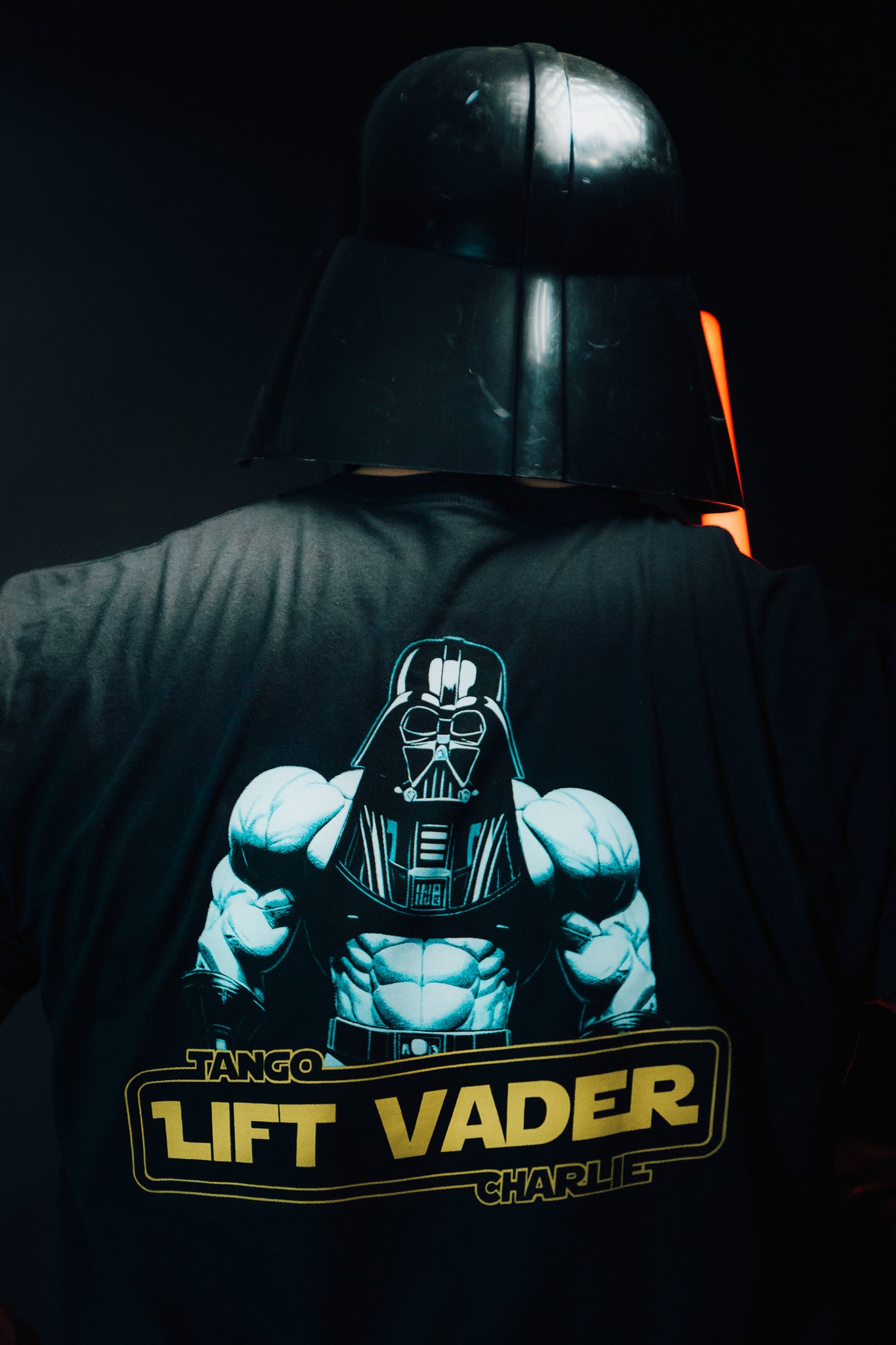 A person wears a helmet and a Tango Charlie Apparels limited edition Lift Vader T-Shirt, displaying a muscular figure with a Darth Vader-like helmet. The dark background is accented with subtle orange light, capturing the essence of fitness and strength.