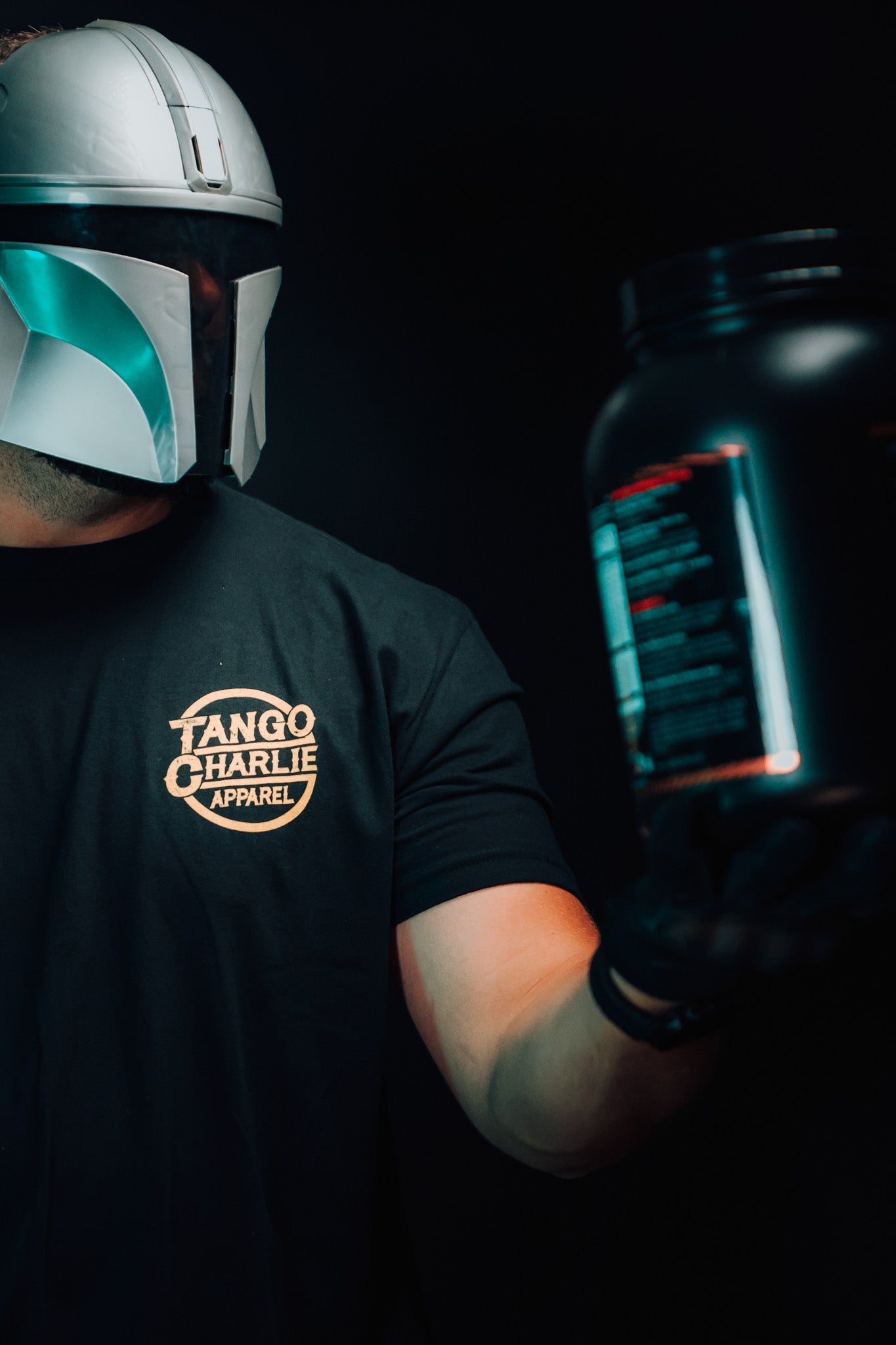 A person in a metallic helmet and a black Tango Charlie Apparel shirt holds up The Whey container against a dark background, highlighting their commitment to muscle building.