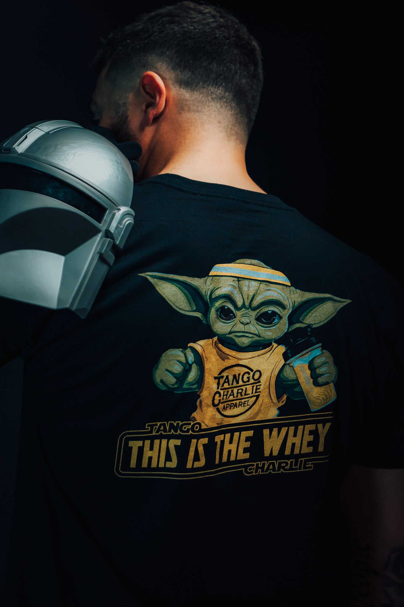 A man with a helmet over his shoulder wears a Tango Charlie Apparel black t-shirt showcasing a green creature with large ears. The shirt, named The Whey, alludes to muscle building and fitness supplements against a dark background.