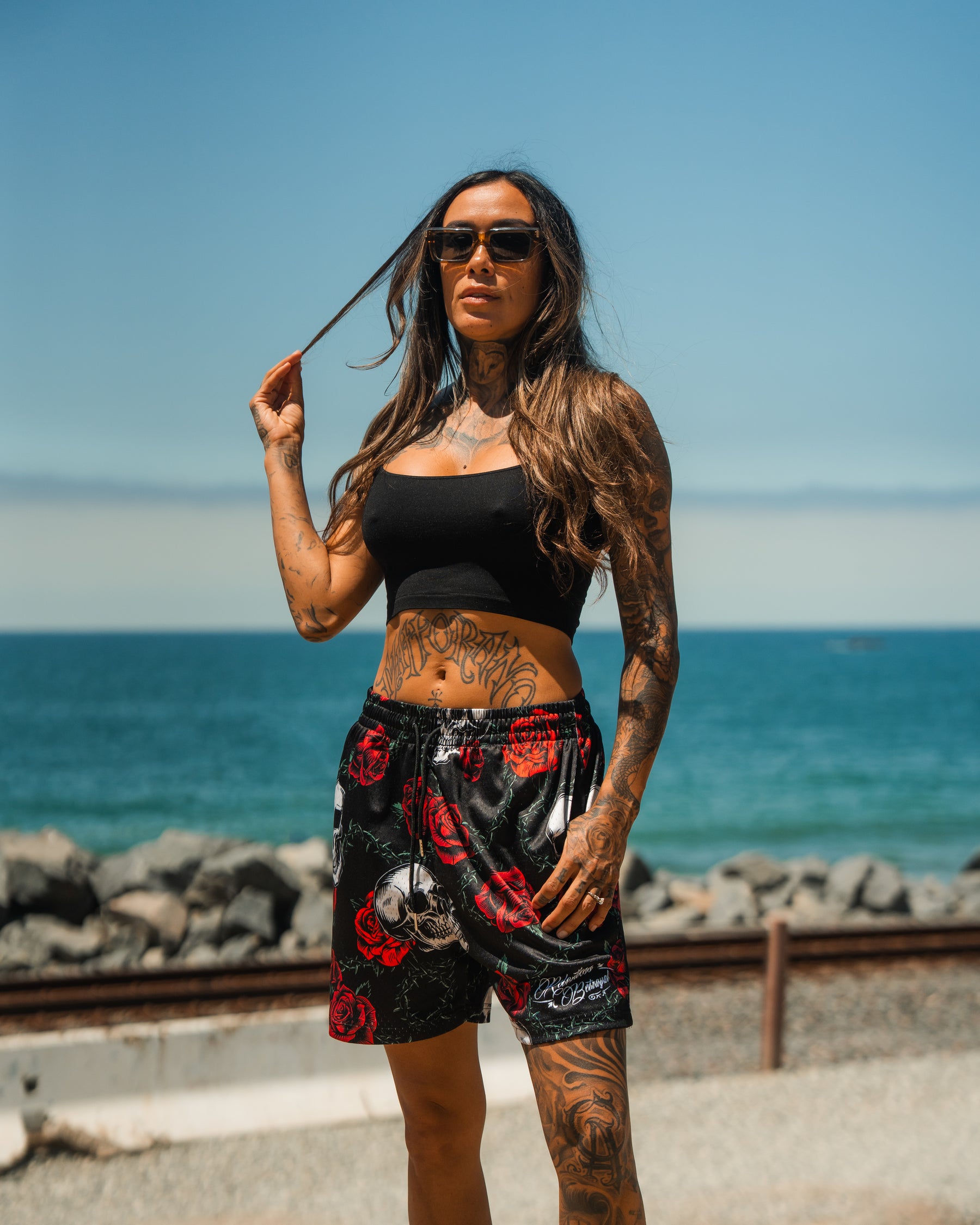A woman with long hair, sunglasses, and tattoos stands by the ocean in a black crop top and Relentless Betrayals Premium Mesh Shorts | 5- Thorn Amongst Roses. Rocks, water, and blue sky form a striking backdrop.