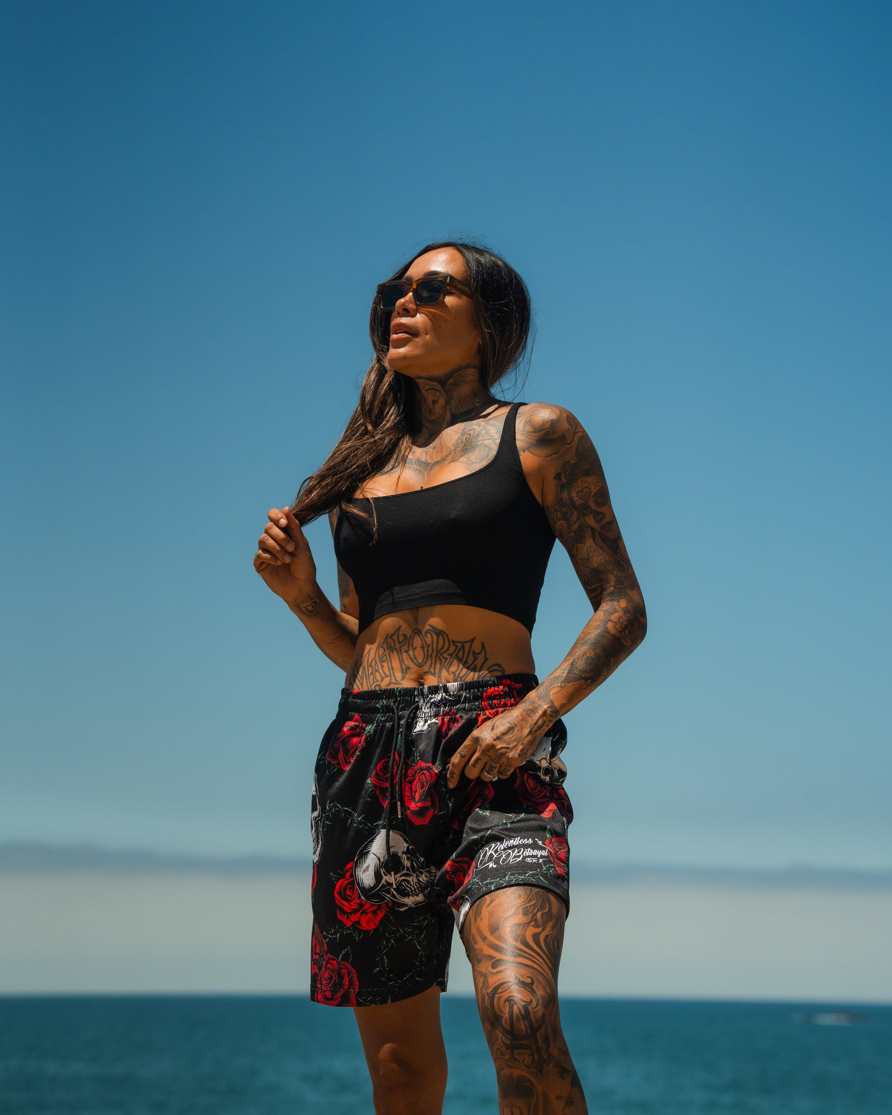 A tattooed woman in sunglasses, a black crop top, and Relentless Betrayals Premium Mesh Shorts | 5- Thorn Amongst Roses stands confidently by the ocean beneath a clear blue sky.