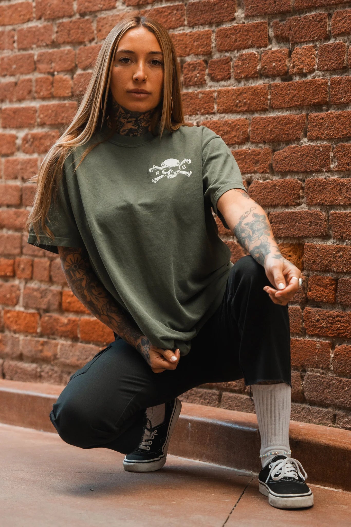 A person with long hair and tattoos crouches against a brick wall, wearing the Relentless Betrayal Tell No Tales Premium Tee in green with a small white graphic, paired with black pants, white socks, and black sneakers.