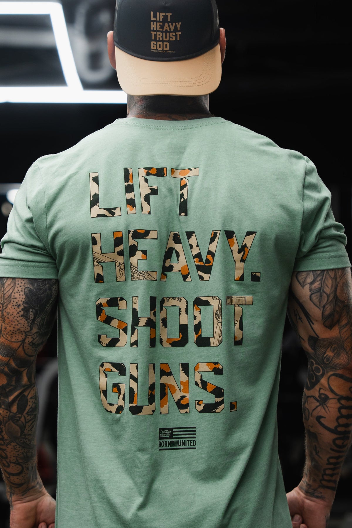 Wearing the Tango Charlie Apparel LHSG - Sage Tee, a person with tattoos stands in a gym filled with heavy lifting and shooting gear, sporting a cap that says "LIFT HEAVY TRUST GOD" and proudly embracing the Second Amendment spirit.