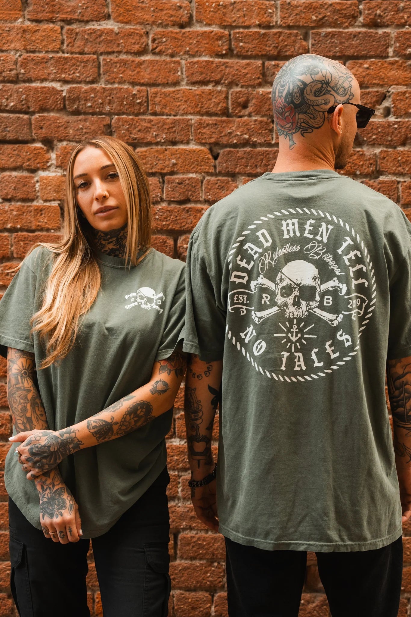 A tattooed woman and man stand before a brick wall; she wears the Relentless Betrayal Tell No Tales Premium Tee- Green with a small skull, while he shows off a similar unisex version featuring Dead Men Tell No Tales and a large skull on the back.