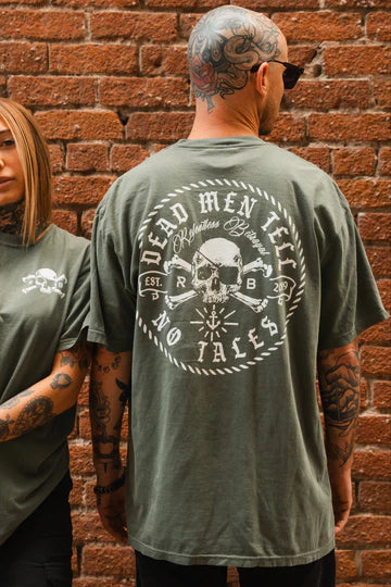 A man in sunglasses leans against a brick wall, flaunting the back of his Tell No Tales Premium Tee by Relentless Betrayal, featuring a skull and Dead Men Tell No Tales. A tattooed woman stands slightly behind him, both showcasing this edgy unisex item.