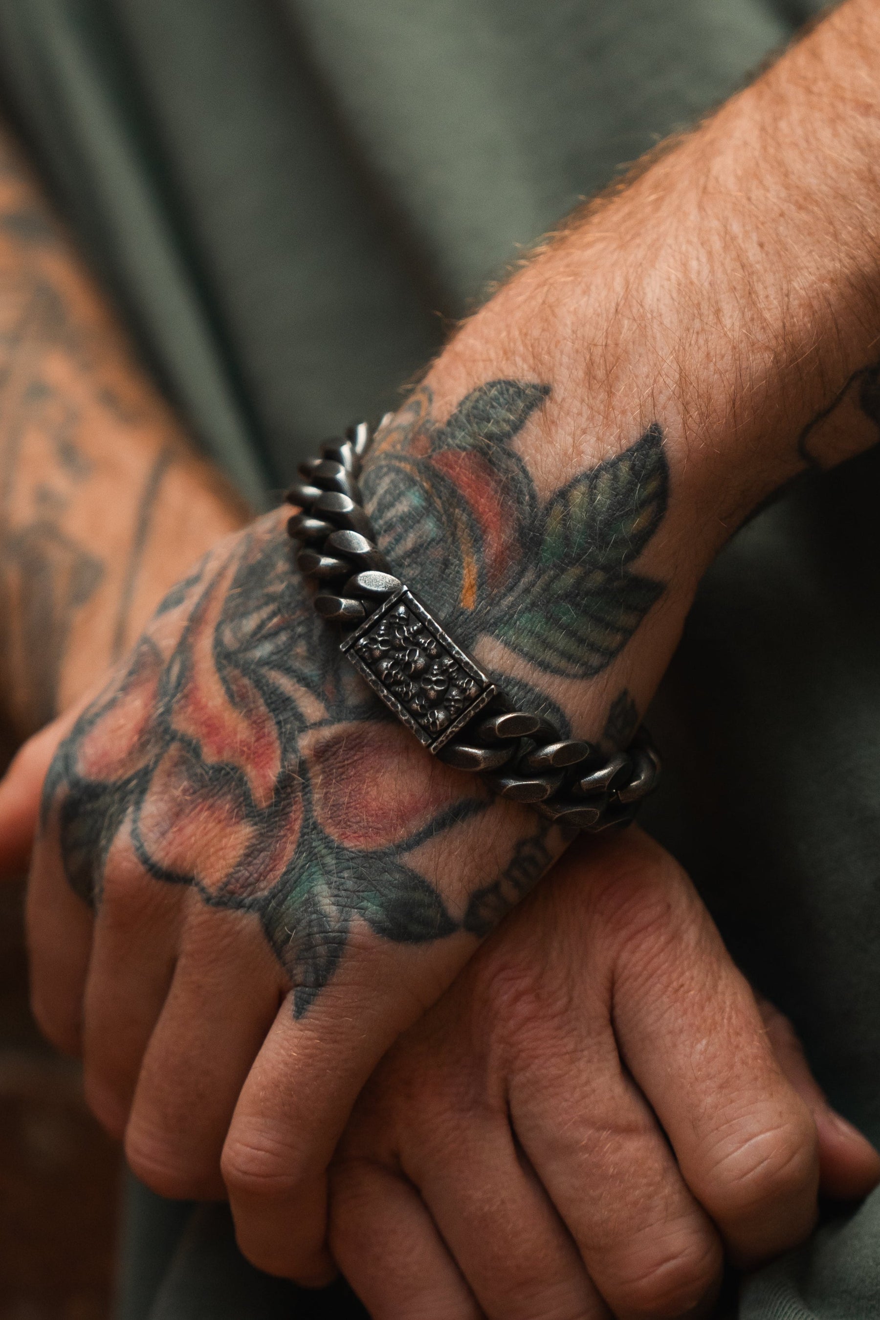 A close-up of a tattooed hand with colorful rose designs resting on fabric, adorned with Relentless Betrayals Tell No Tales chunky stainless steel bracelet, featuring an ornate rectangular design. The person is wearing a green shirt.