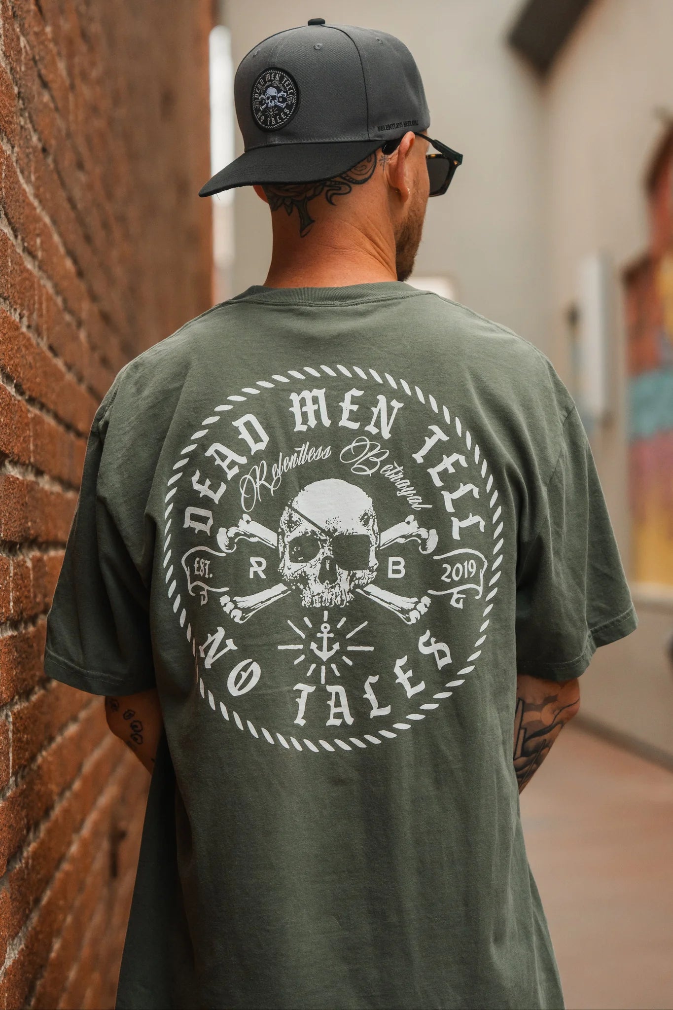 In an alleyway, a man wears Relentless Betrayals Tell No Tales Premium Tee in green, featuring a skull and crossbones with Dead Men Tell No Tales. Paired with tattoos, a gray cap, and sunglasses, his style is edgy yet effortlessly cool.