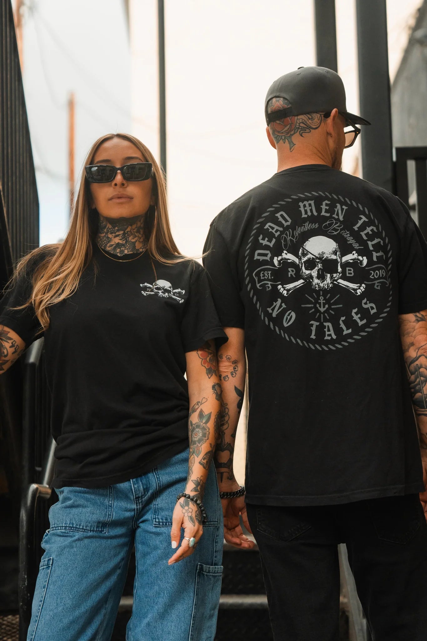 A person with long hair and sunglasses wears Relentless Betrayals Tell No Tales Premium Tee, featuring a skull design, standing next to someone in a black cap and a shirt with Dead Men Tell No Tales and skulls on the back. Both have tattoos.