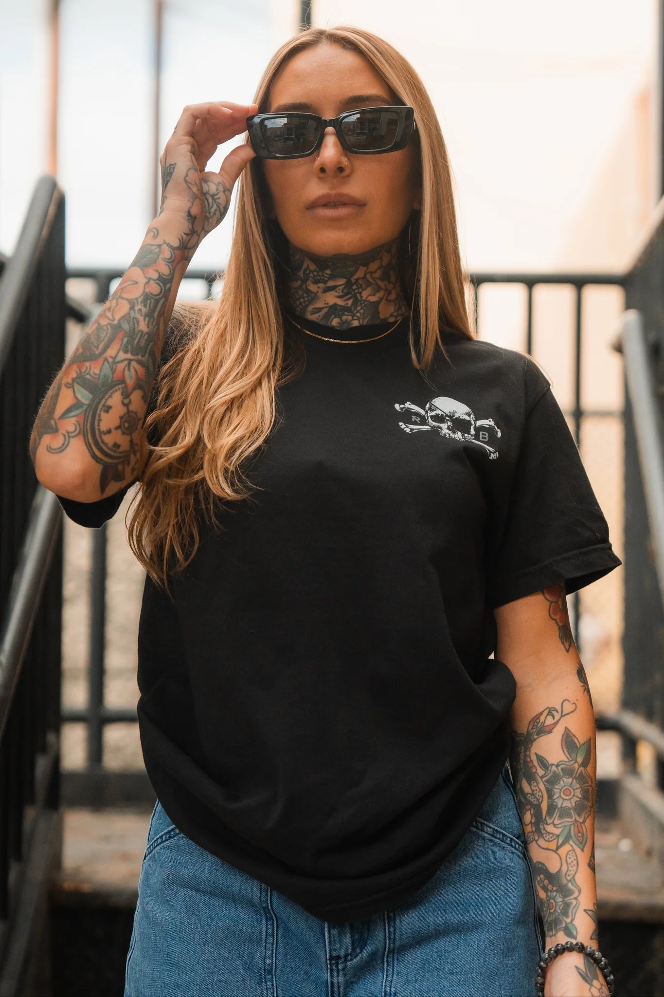 A confident individual with long hair and tattooed arms and neck stands wearing sunglasses, blue jeans, and the Relentless Betrayal Tell No Tales Premium Tee, a black jersey cotton t-shirt with a small white skull design. The background features a staircase and railing.