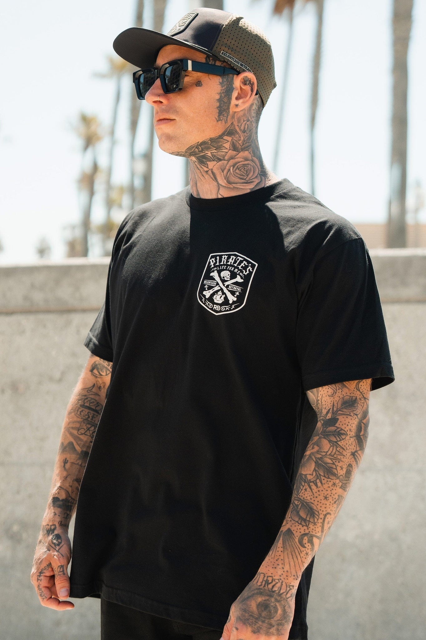 A tattooed person in sunglasses, a cap, and a Relentless Betrayal Pirates Life Black T-Shirt stands outdoors with palm trees and a concrete wall in the background under a clear sky, exuding adventure.