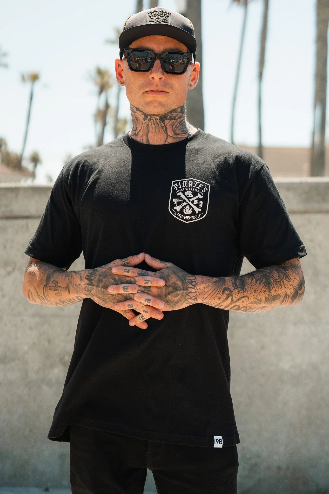 A person in a Relentless Betrayal Pirates Life Black T-Shirt, sunglasses, and a black baseball cap stands confidently outdoors. Tattooed arms add to their style against a backdrop of palm trees and a concrete wall on a sunny day.
