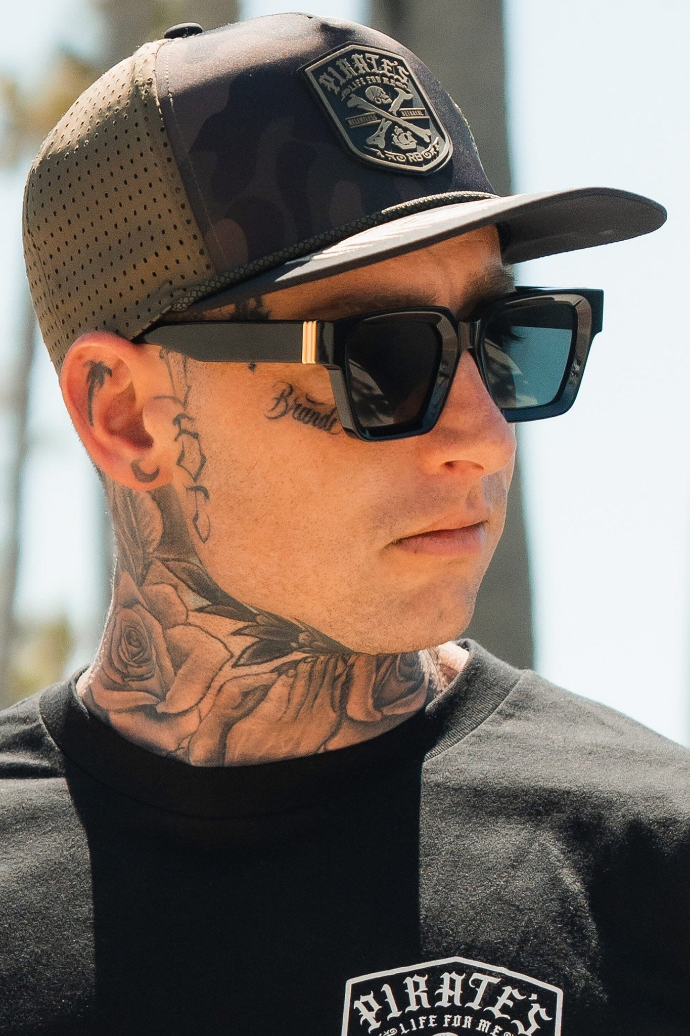 Wearing a Relentless Betrayal Pirates Life Camo Snapback, sunglasses, and a black Pirates T-shirt, a tattooed man exudes adventure. An intriguing tattoo under his eye and blurred tree or pole in the background complete the scene.