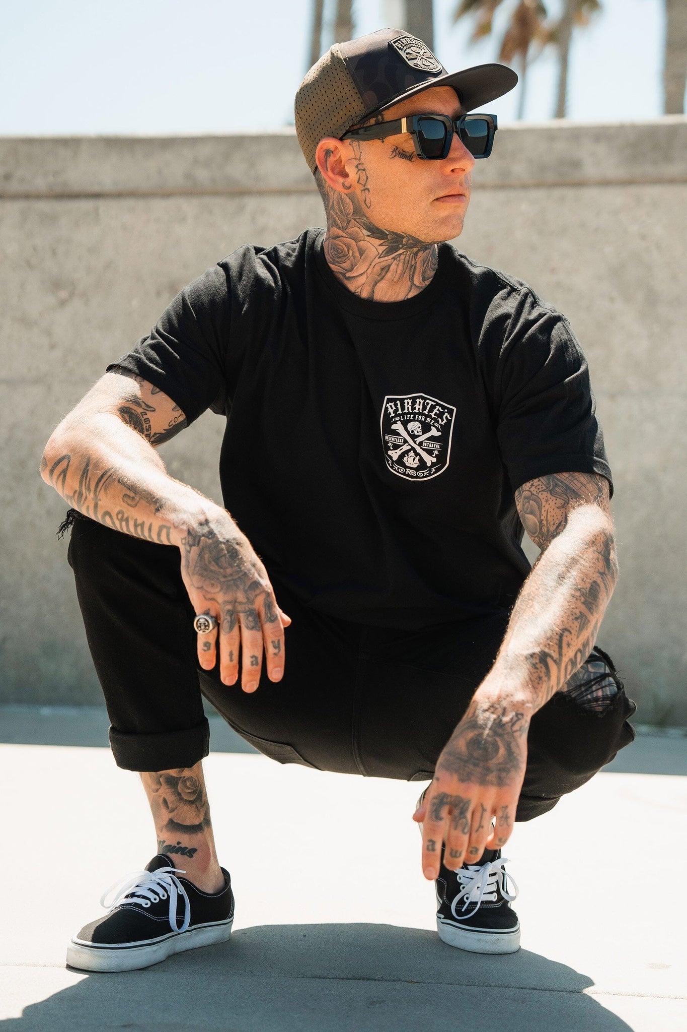 A tattooed person squats on a sunny street, wearing a black cap, sunglasses, and a Relentless Betrayal Pirates Life Black T-Shirt. Theyre dressed in all-black with pants and sneakers against palm trees and a concrete wall.