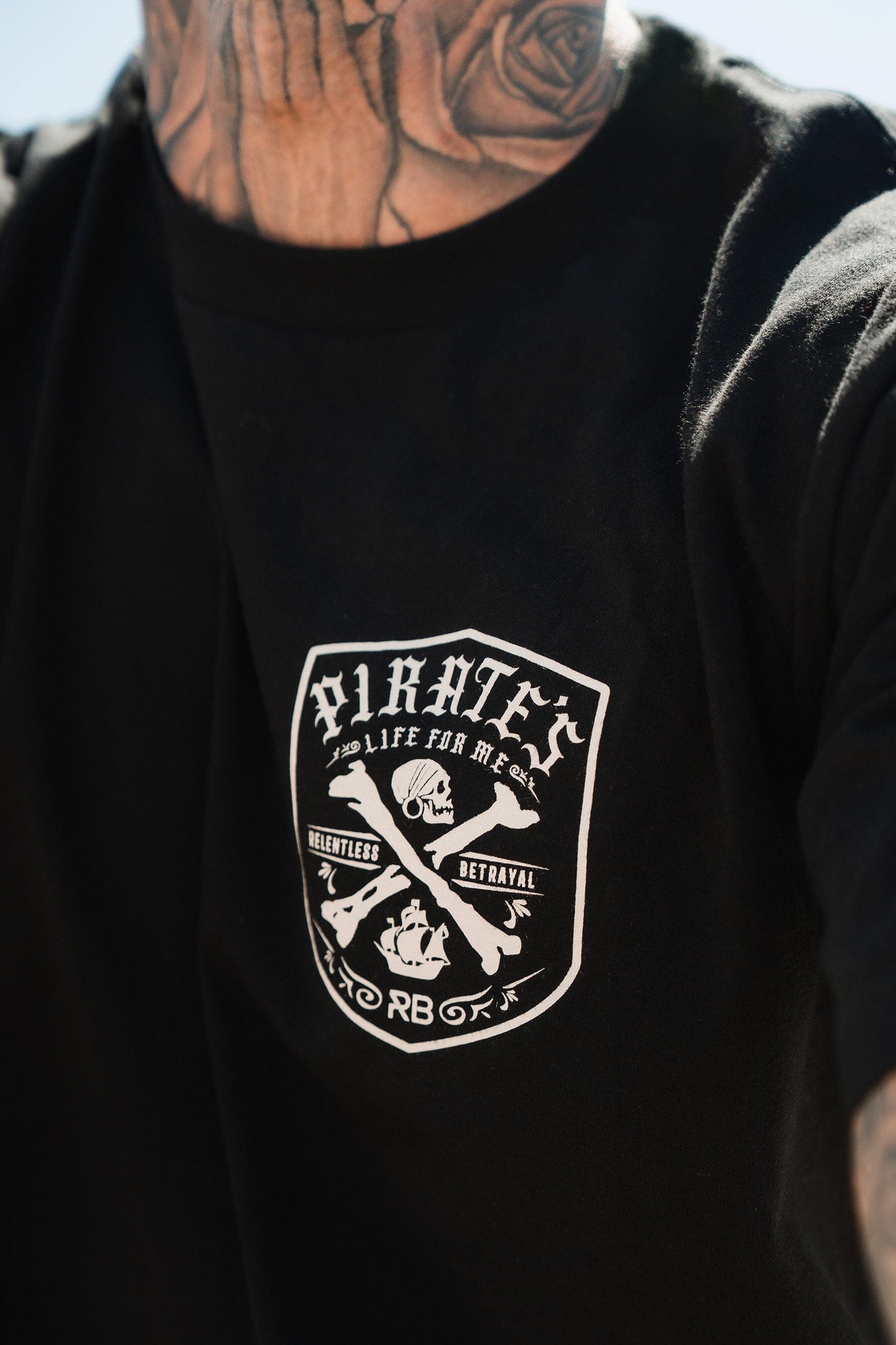A close-up shows someone wearing the Pirates Life Black T-Shirt by Relentless Betrayal, featuring a pirate-themed emblem with crossed bones, ships wheel, and skull alongside the bold phrases Pirates Life, Relentless Adventure, and Betrayal on black fabric.