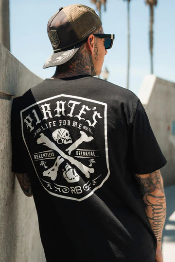 A tattooed man in a baseball cap and sunglasses strikes an adventurous pose outdoors, wearing Relentless Betrayals Pirates Life Black T-Shirt with a bold skull and crossbones design. Swaying palm trees complete his daring look.