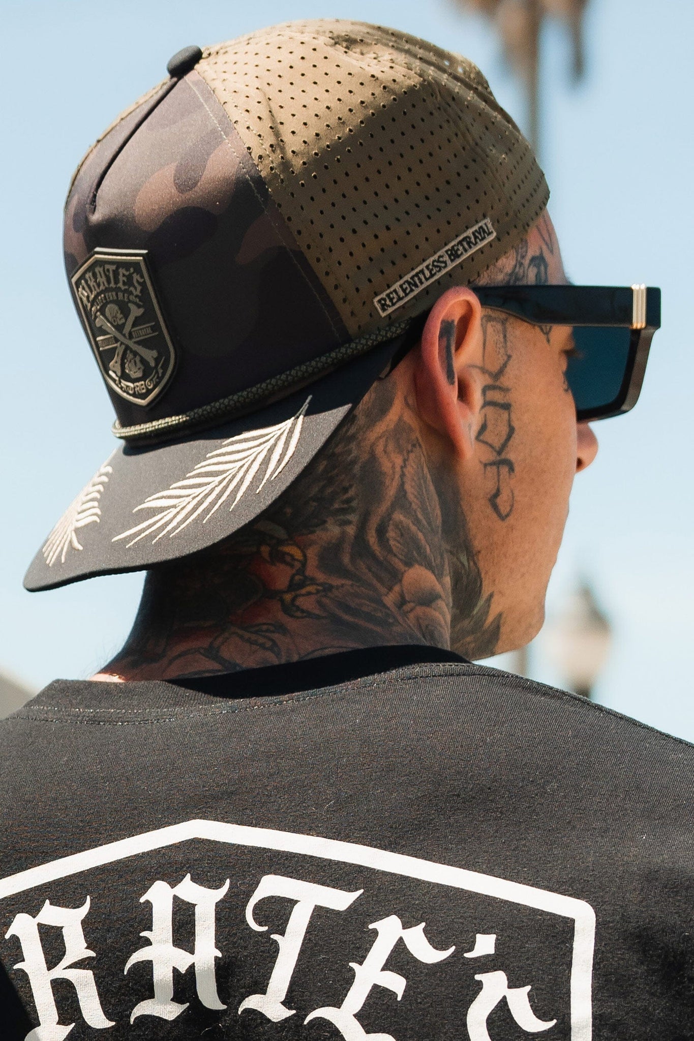 A person with neck tattoos wears a Pirates Life Camo Snapback by Relentless Betrayal, featuring a patch and RELENTLESS BETRAYAL on the strap. They have large sunglasses, a black shirt with bold white text, exuding adventure while keeping their face hidden.