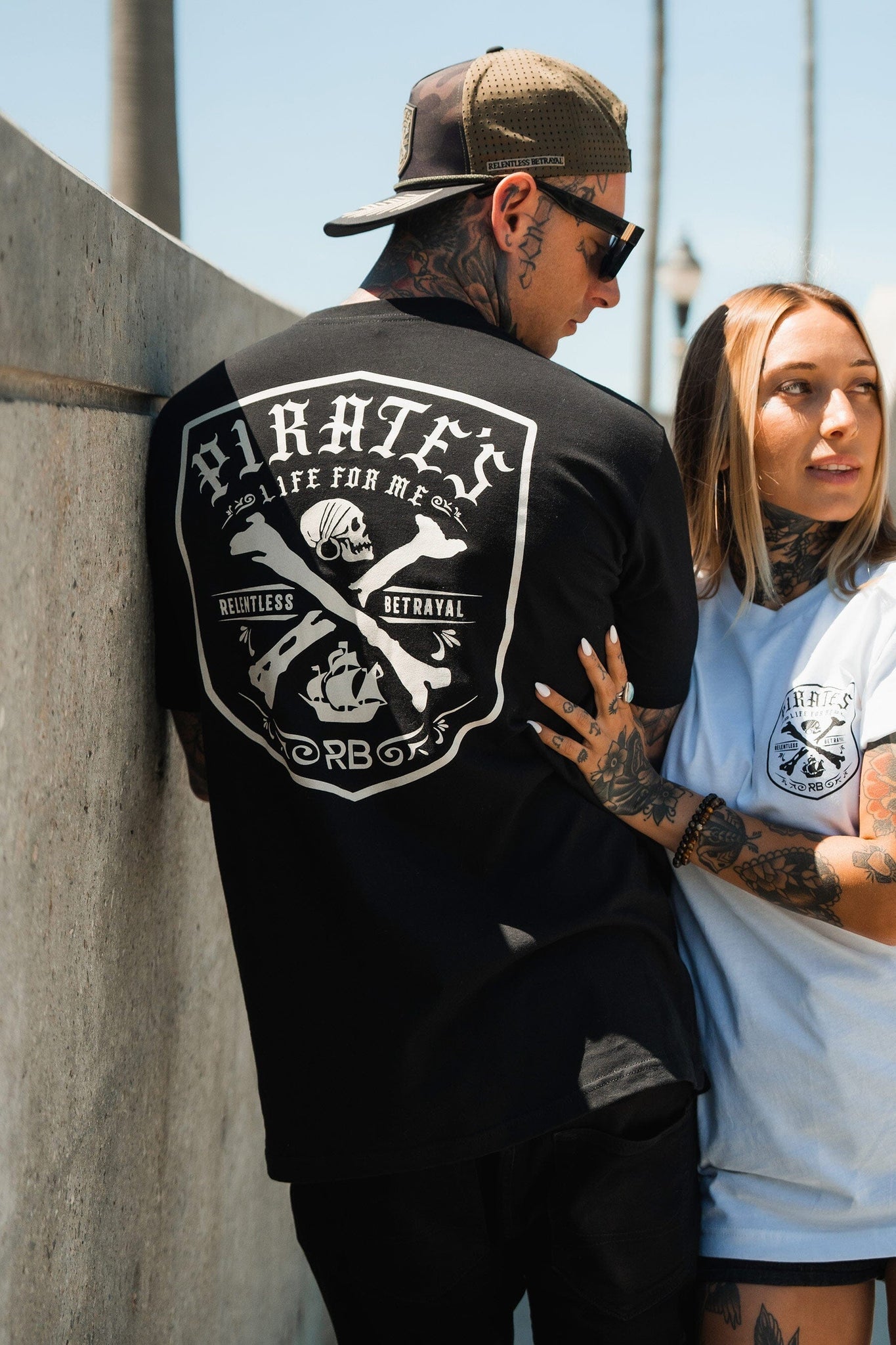 A tattooed couple stands close together under the sunny sky. The man sports a Relentless Betrayal Pirates Life Black T-Shirt with a graphic on the back and a cap, while the woman wears a white shirt echoing the adventurous design on her chest.