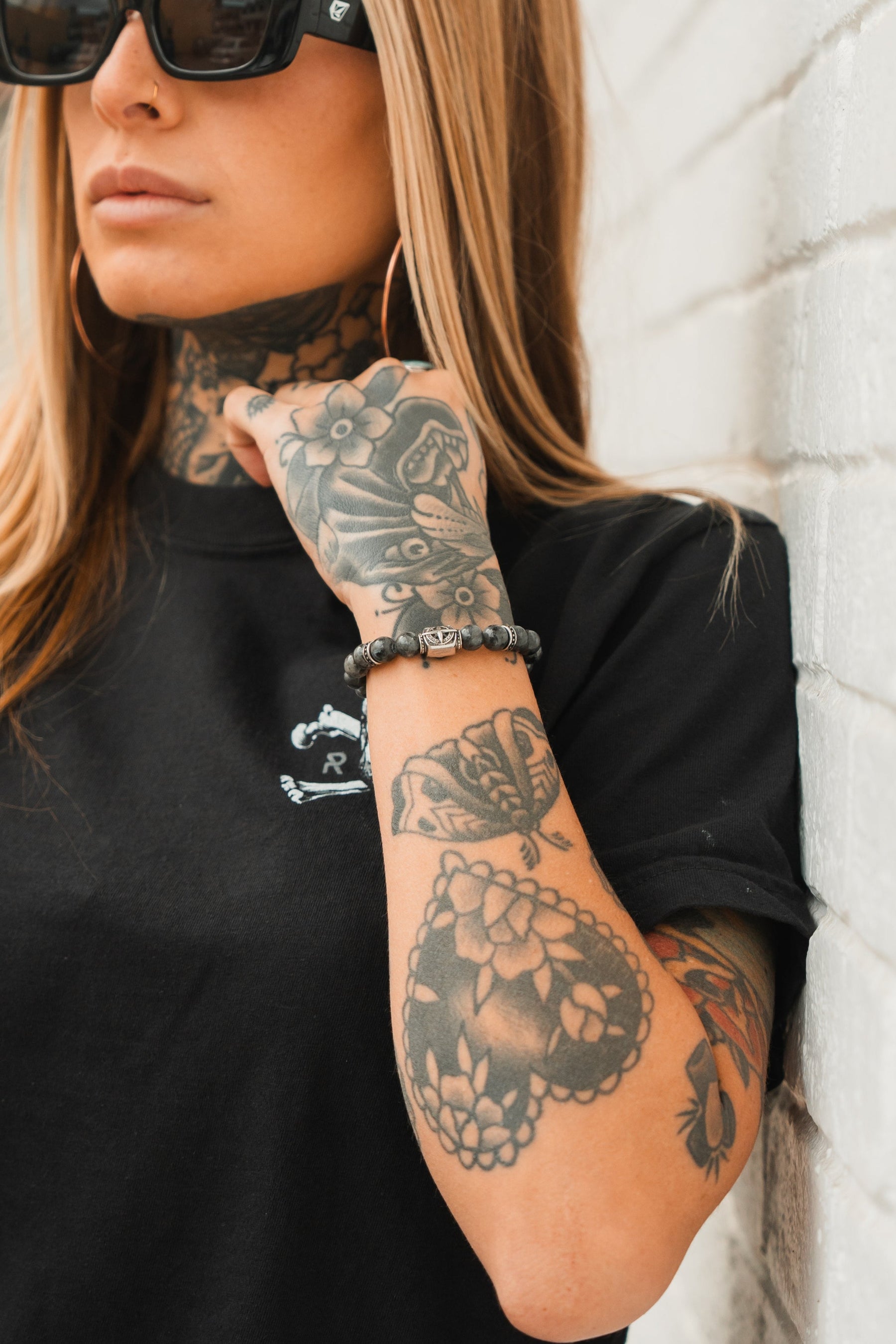 A person with blonde hair and sunglasses leans against a white brick wall, flaunting neck, hand, and arm tattoos of floral and abstract designs. They wear a black t-shirt with a Polaris Bead Bracelet from Relentless Betrayal, adding an intriguing touch.