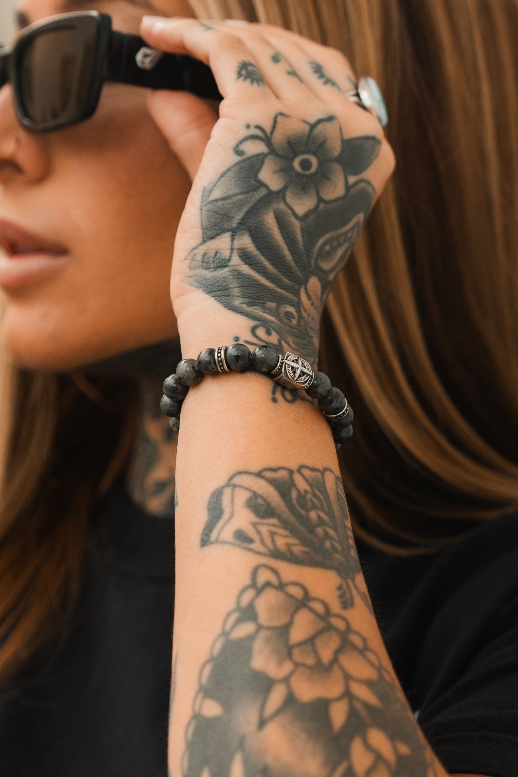 A person with long brown hair and black sunglasses showcases a heavily tattooed floral arm. Theyre wearing a dark shirt, a Polaris Necklace with sparkling gemstones and star pendant, complemented by a beaded bracelet from Relentless Betrayal named the Polaris Bead Bracelet.