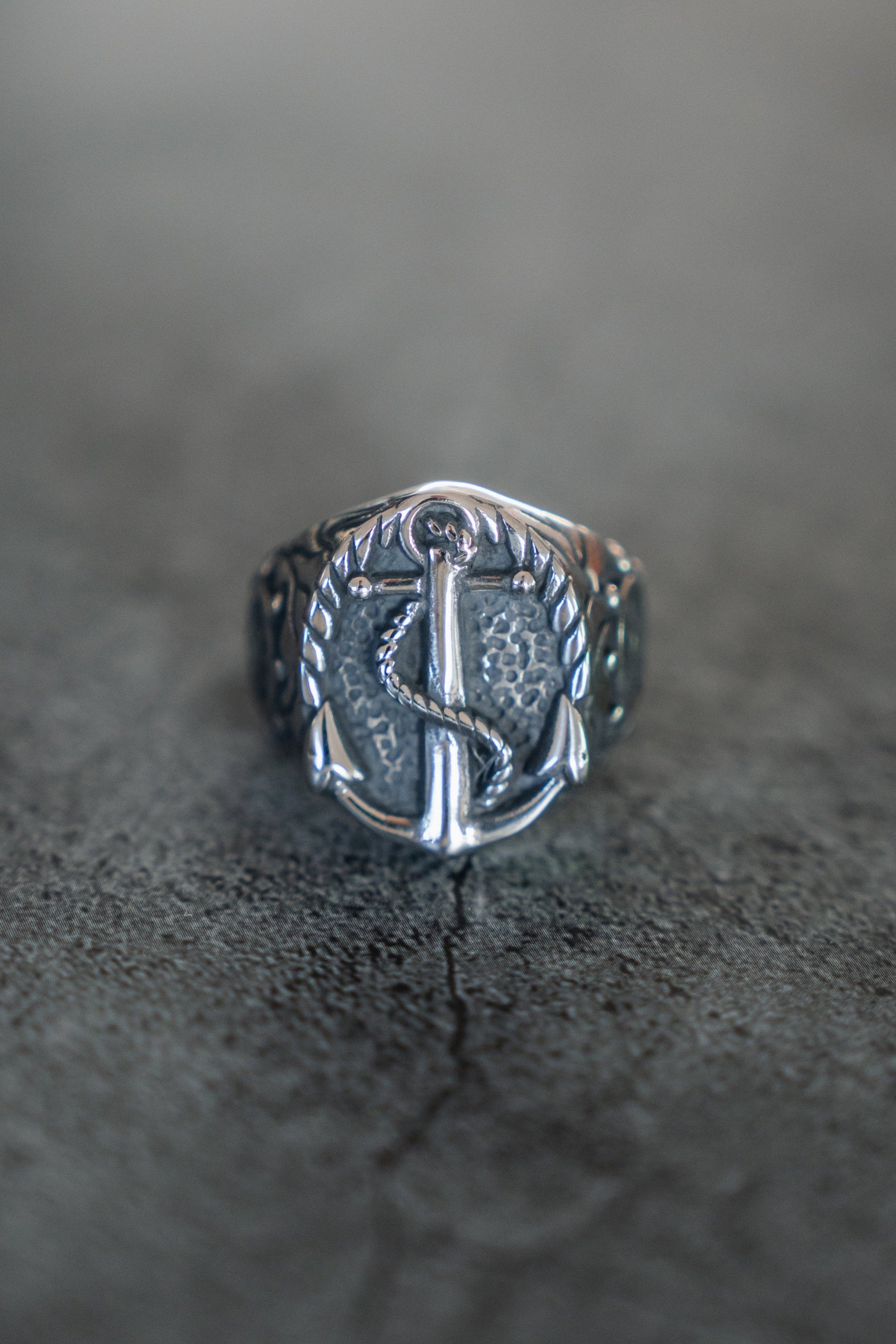 Discover the Voyager Ring by Relentless Betrayal: a stainless steel ring with an engraved anchor and wreath-like pattern, ideal for adventure enthusiasts. This silver masterpiece is showcased on a textured gray surface.