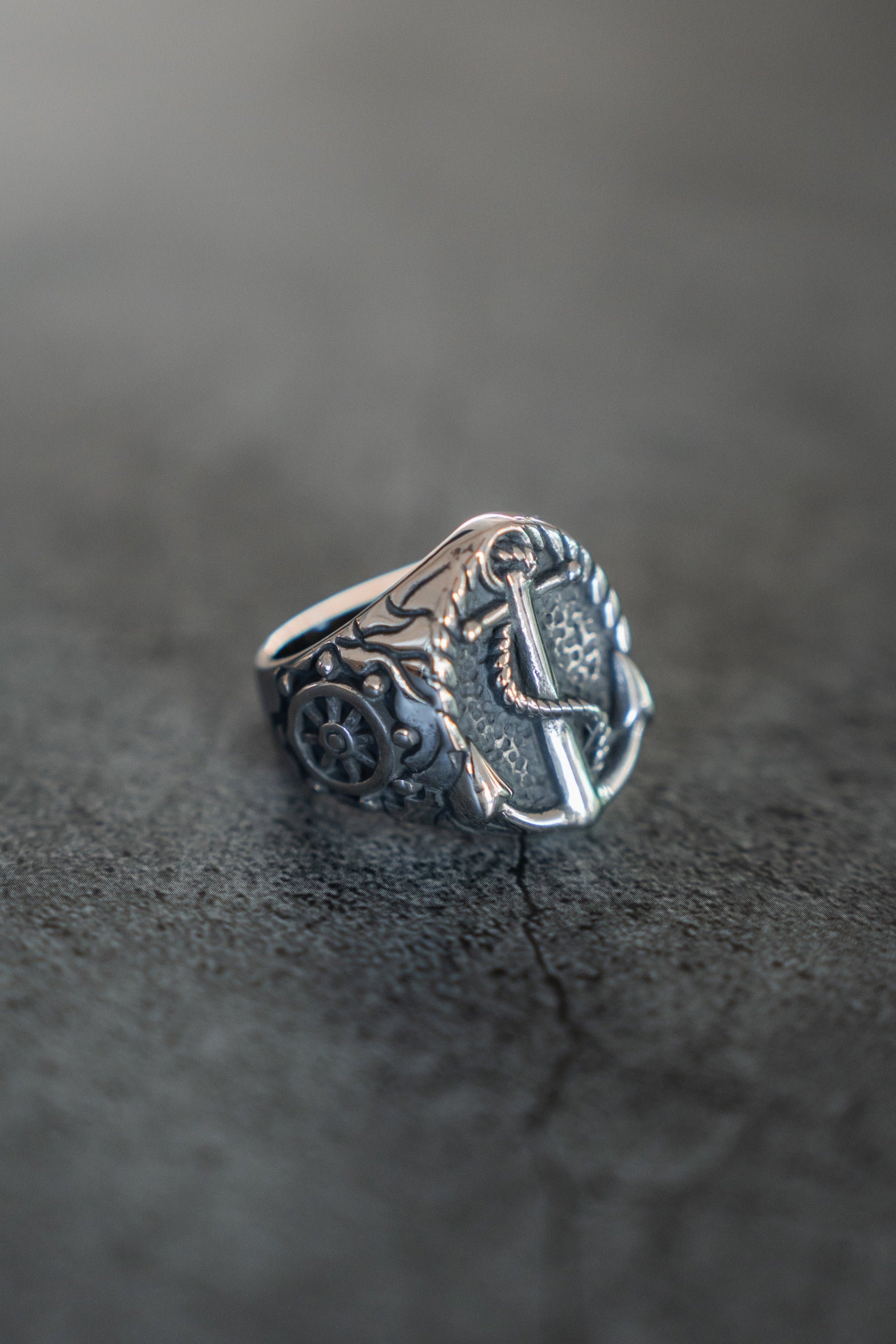 Introducing the Voyager Ring by Relentless Betrayal: a silver band with tree engravings on a textured gray surface, inspired by adventure jewelry. It features a detailed round floral pattern on the side, showcasing exquisite craftsmanship.