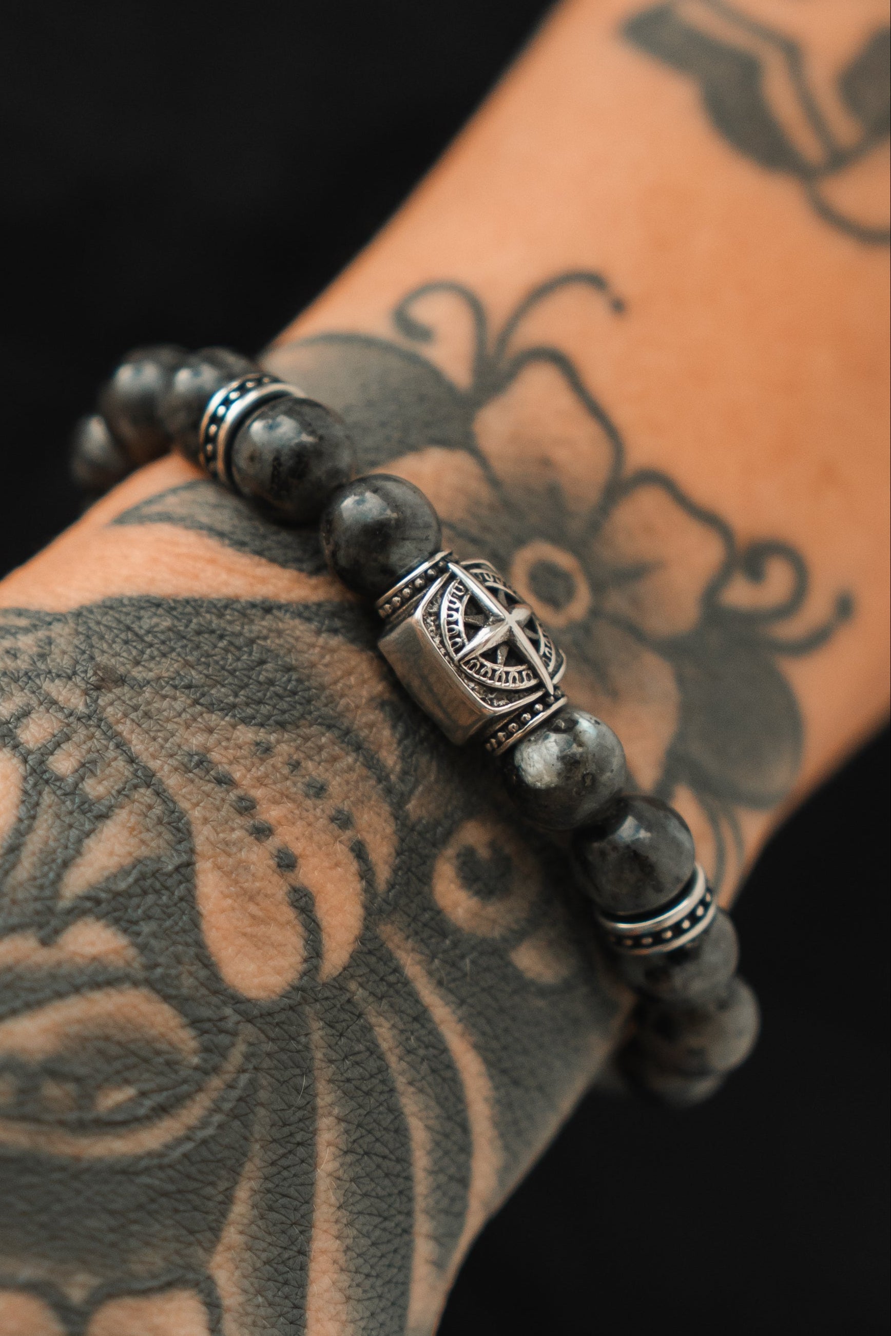 A wrist with intricate tattoos wears the Relentless Betrayal Polaris Bead Bracelet. Featuring gray and black stones and a central silver star-shaped pendant, it strikingly contrasts with the detailed floral and abstract tattoo patterns on the skin.