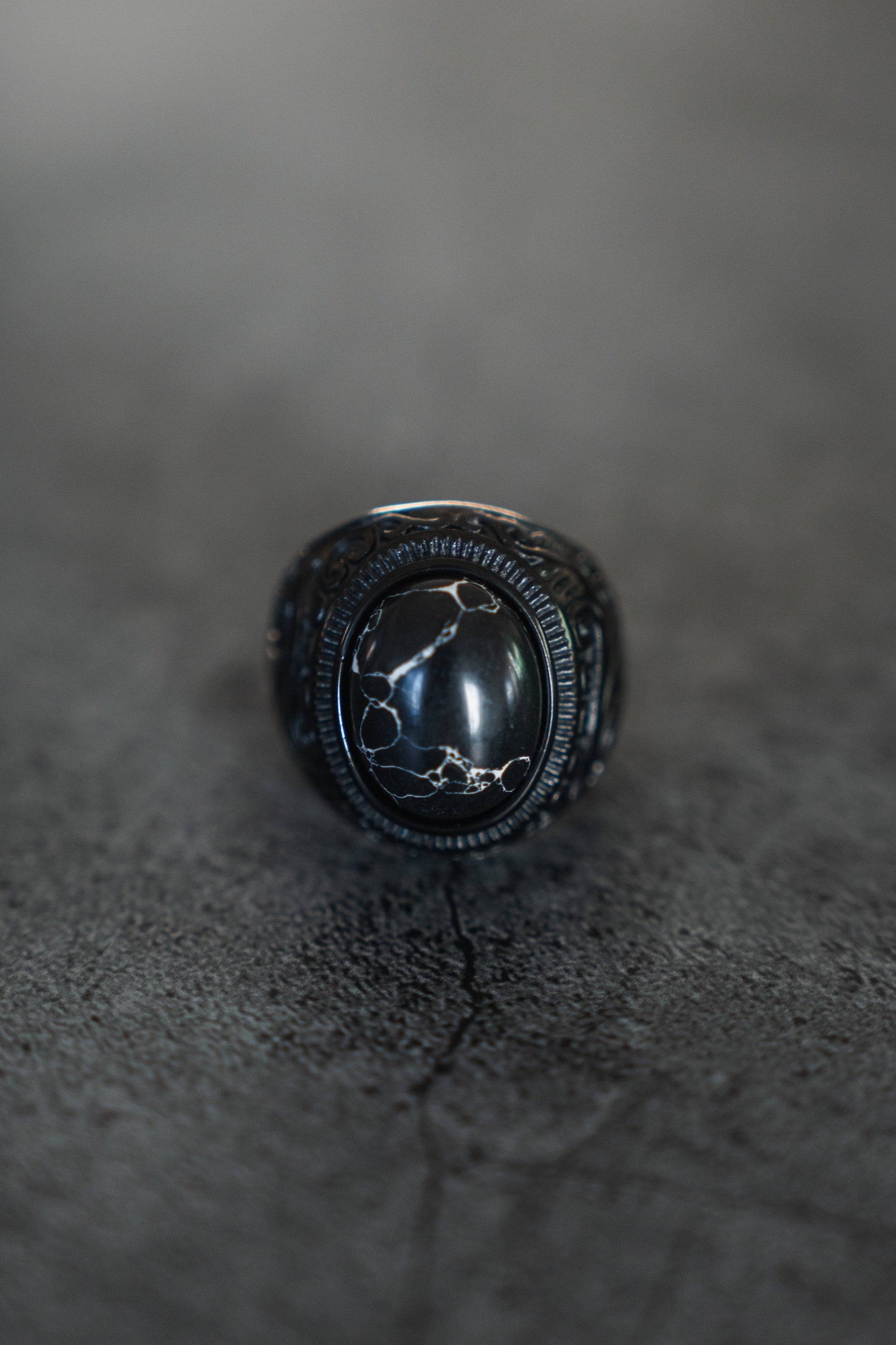 A close-up of Relentless Betrayals The Dark Storm Ring displays an oval black stone with white veining patterns set in an ornate stainless steel band, resting on a textured gray surface for a striking contrast.